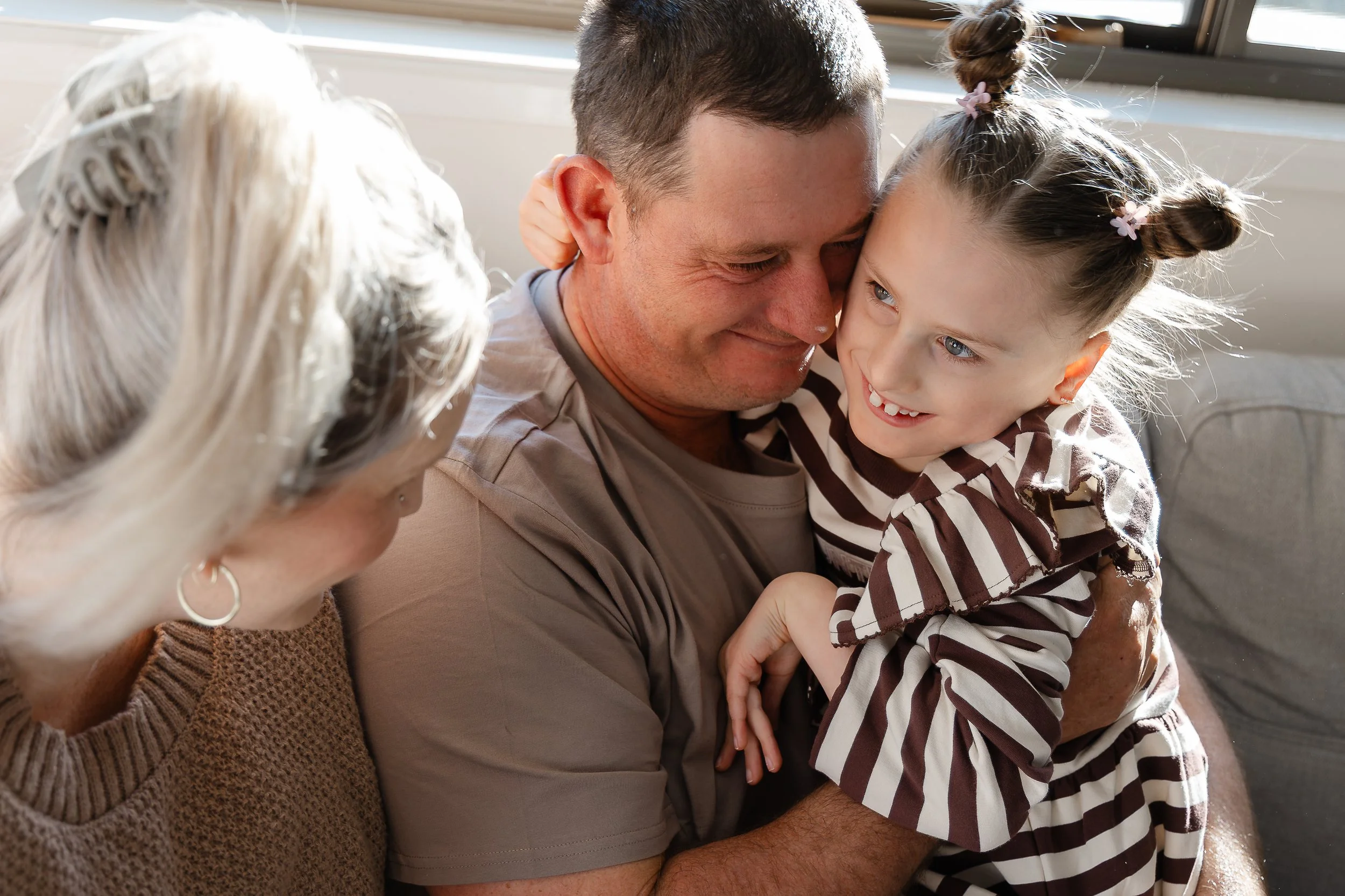 A young girl and a man, likely her father, are close together indoors, smiling and touching foreheads, with a woman with white hair partially visible in the foreground, all sharing a warm moment.