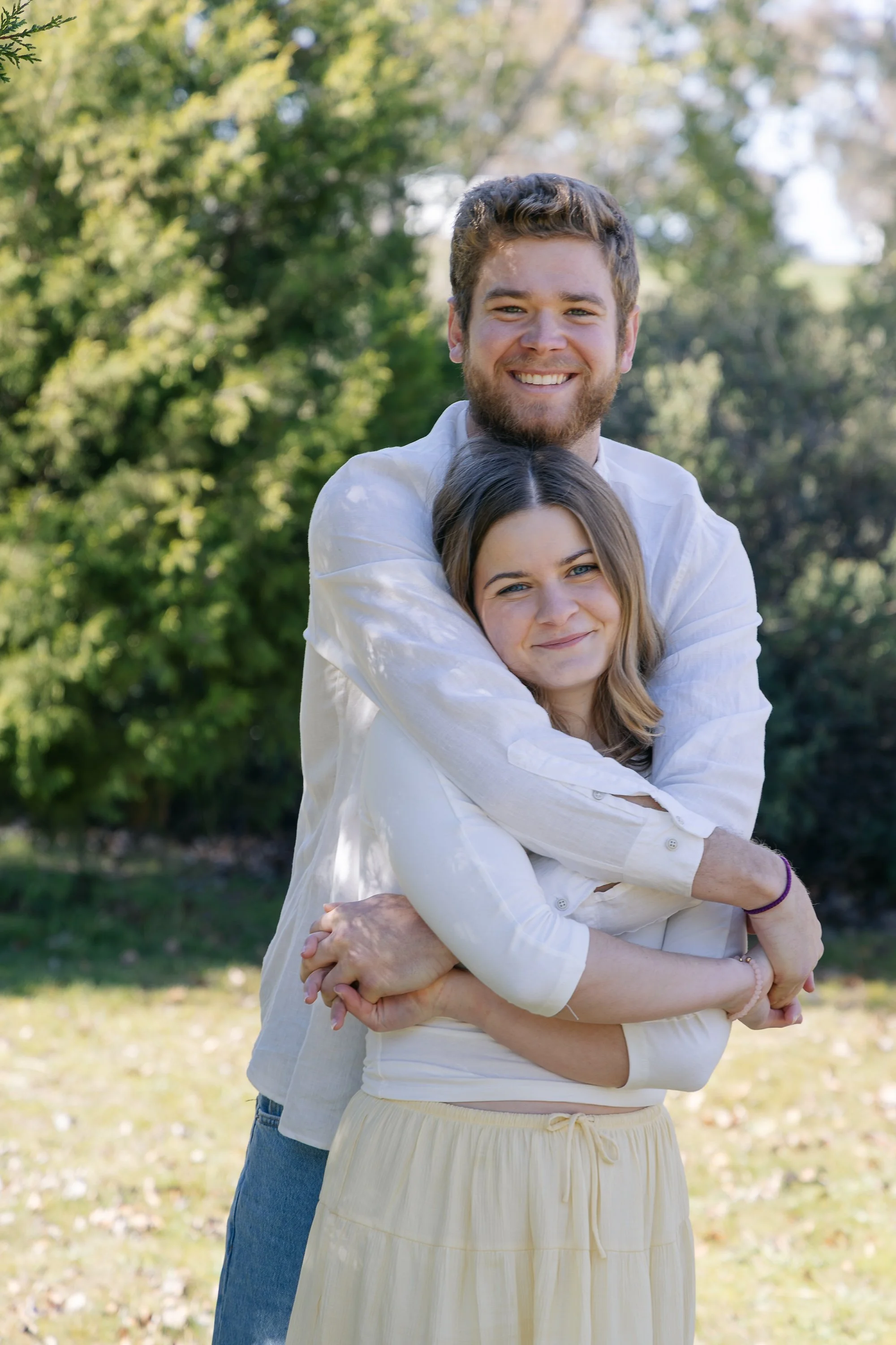 A young man with a beard and a young woman with long brown hair are smiling and hugging outdoors on a sunny day, with green trees in the background.