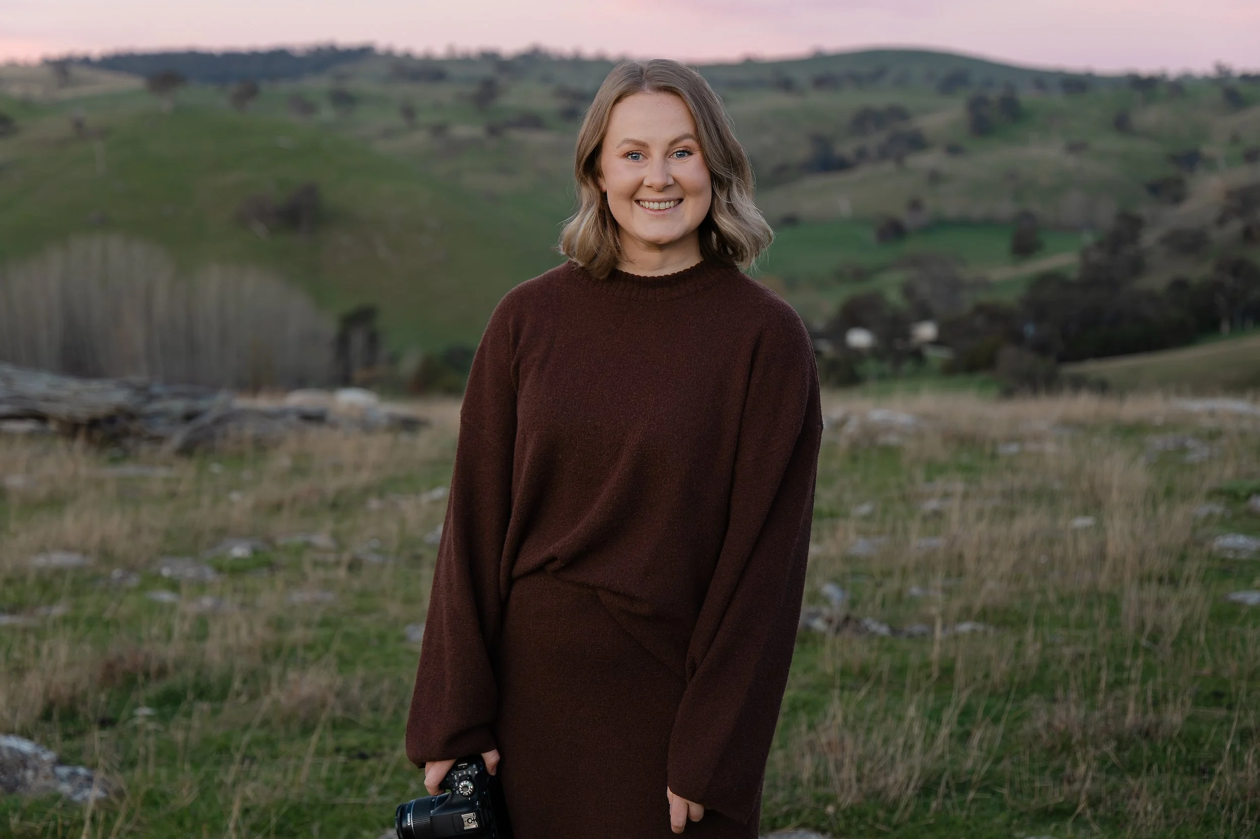 A woman with shoulder-length brown hair smiling outdoors during sunset, holding a camera in her right hand, with a hilly landscape and trees in the background.