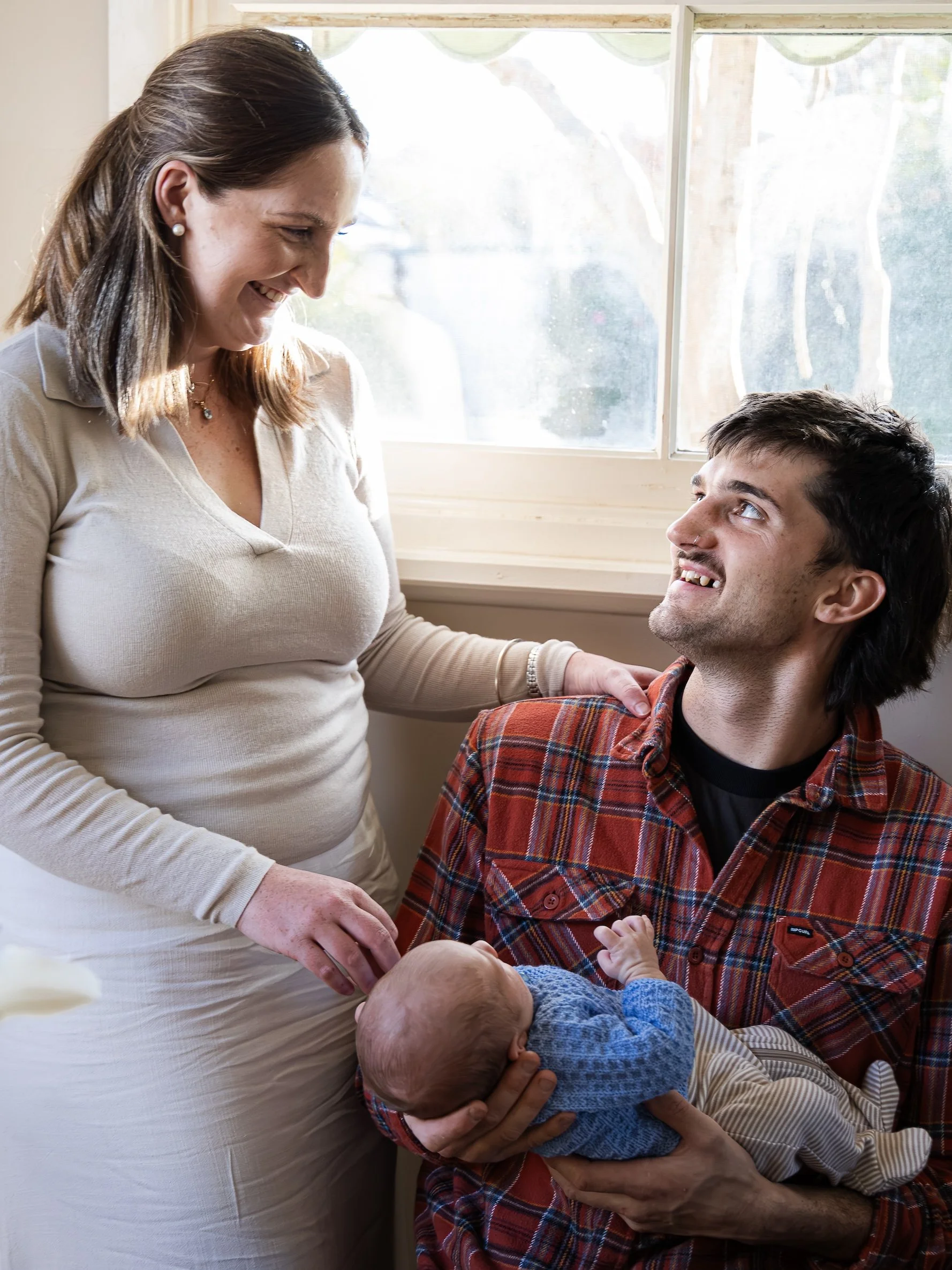 A woman with brown hair and pearl earrings smiling at a man with dark hair and a beard, sitting and holding a baby in an indoor setting near a window.