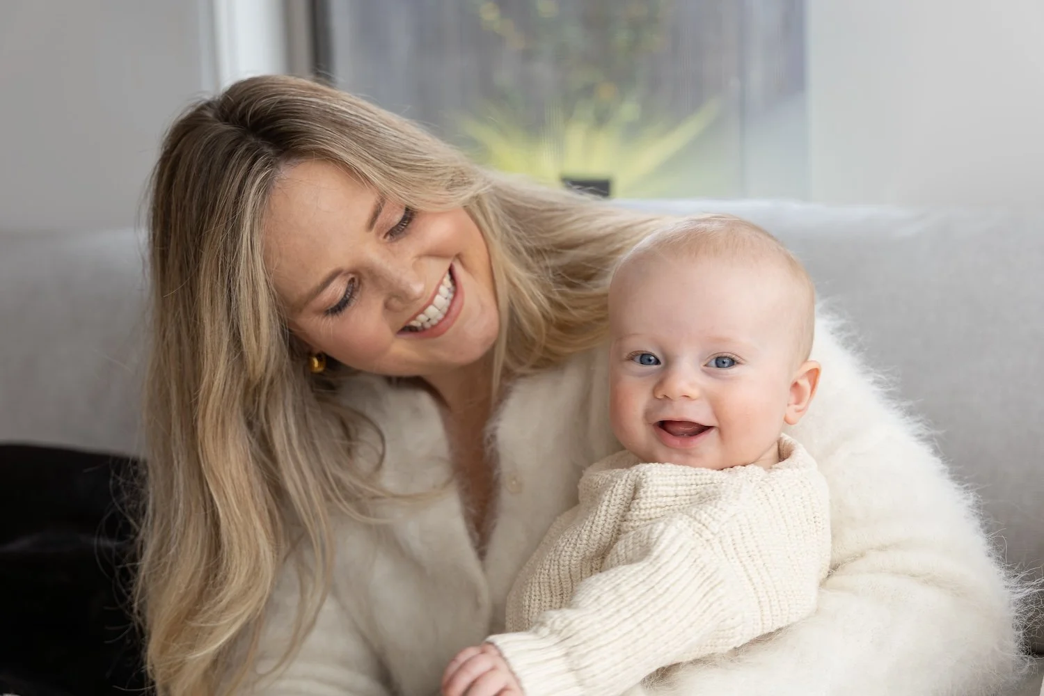 A woman with blonde hair and a white sweater smiling while holding a baby with blue eyes and a beige sweater on her lap in a cozy, well-lit room.