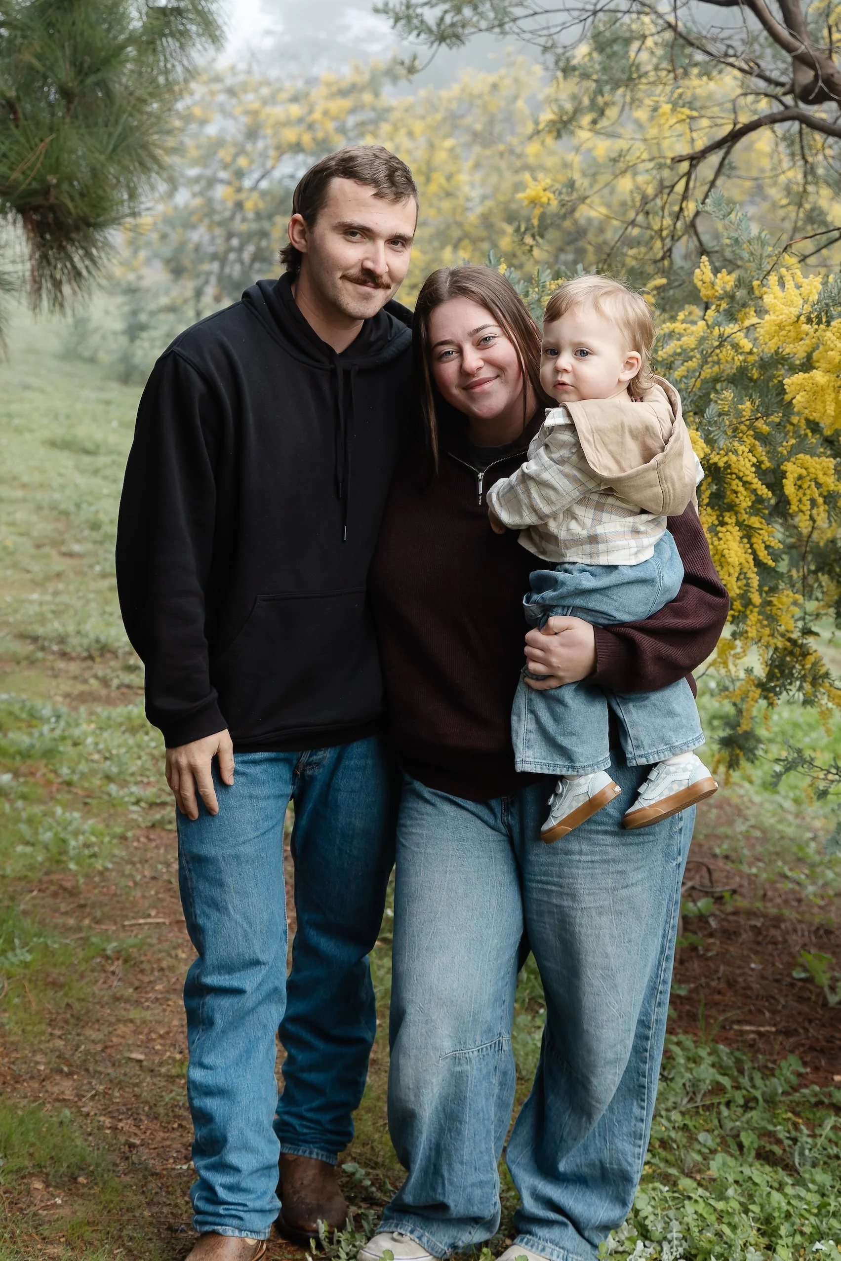 A family of three standing outdoors on a trail with trees and yellow flowers in the background. The man has a mustache and is wearing a black hoodie, the woman has long dark hair and is wearing a dark sweater, and the child is being held by the woman