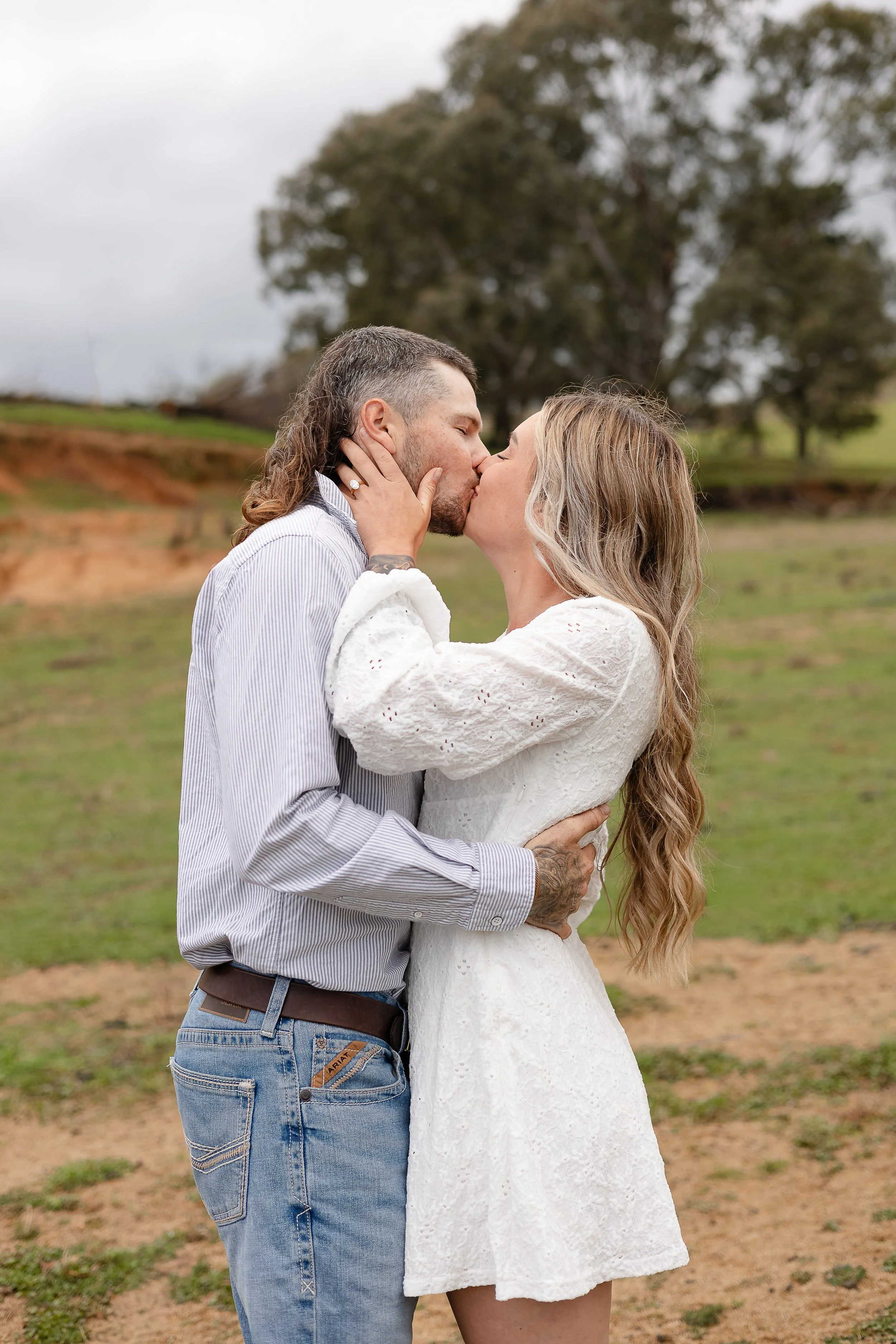Natural engagement photography in Yass, NSW, capturing a genuine moment as a couple share a kiss outdoors. The bride-to-be wears a beautiful white dress with long, softly curled blonde hair, while her partner wears jeans and a tucked-in button-up shi