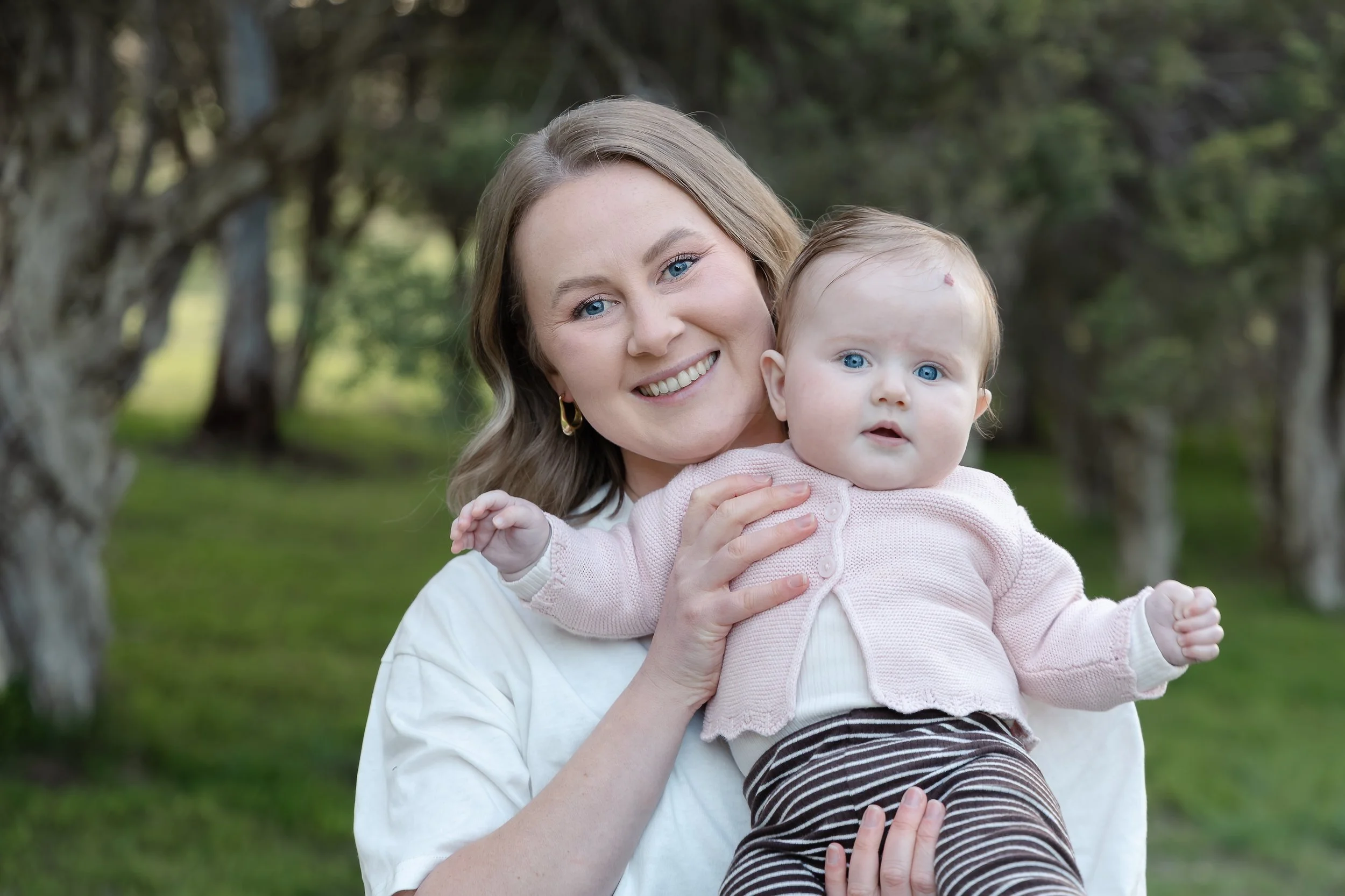 A woman with blonde hair and blue eyes smiling while holding a baby girl outdoors in a park with trees in the background.