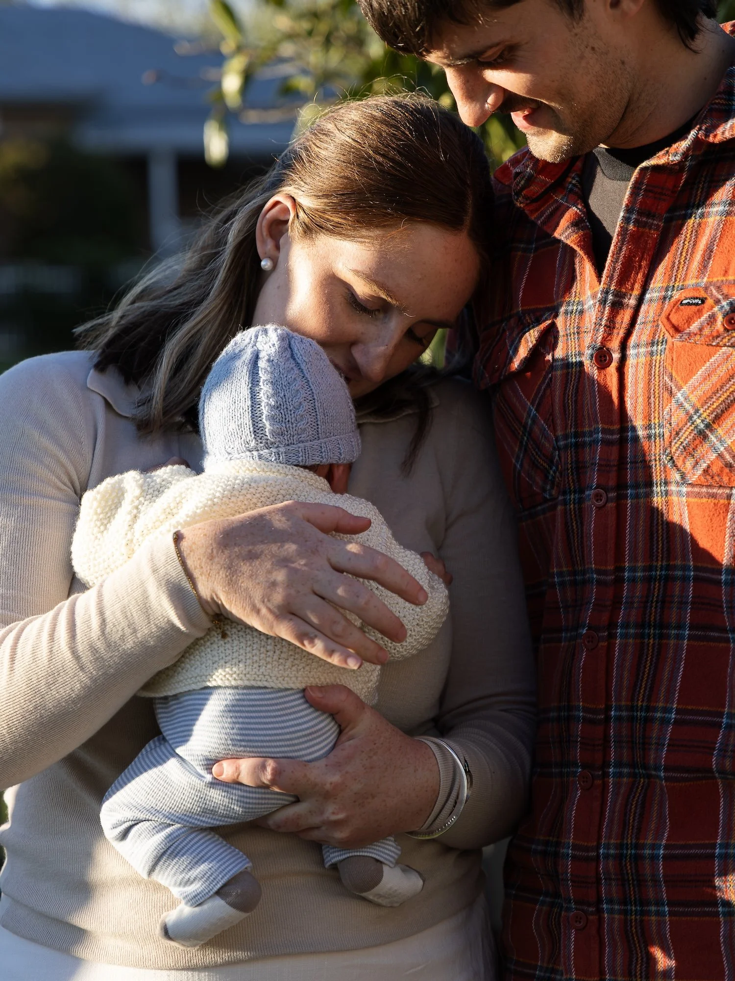 A woman holding a baby close to her chest, with a man standing beside them. The woman has her eyes closed, smiling softly, and is wearing a beige sweater and earrings. The man is wearing a red plaid shirt and is leaning close to the woman. The settin