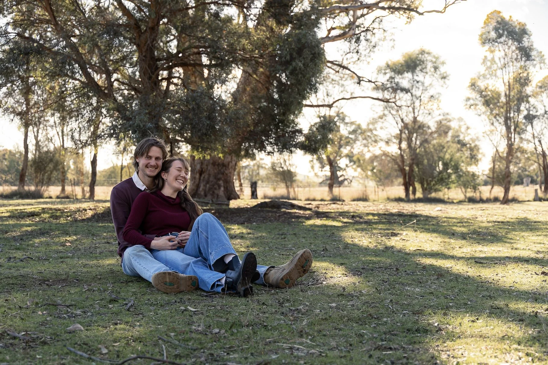 A young couple sitting on the grass in a park, smiling and enjoying each other's company during a sunny day. The background features large trees and bright sunlight filtering through the leaves.
