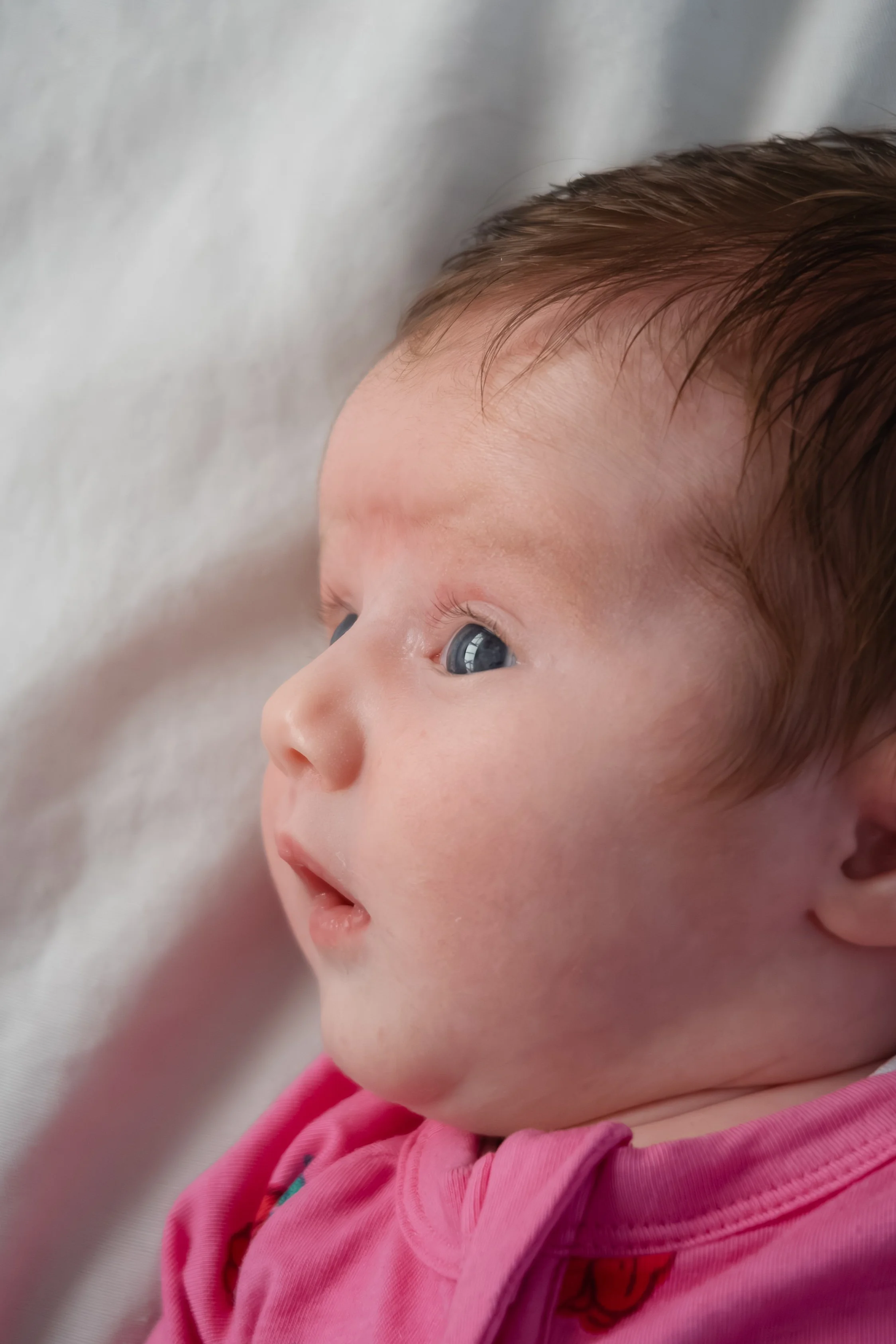 Close-up of a baby's face showing blue eyes, reddish-brown hair, and wearing a pink outfit with a small embroidered detail.