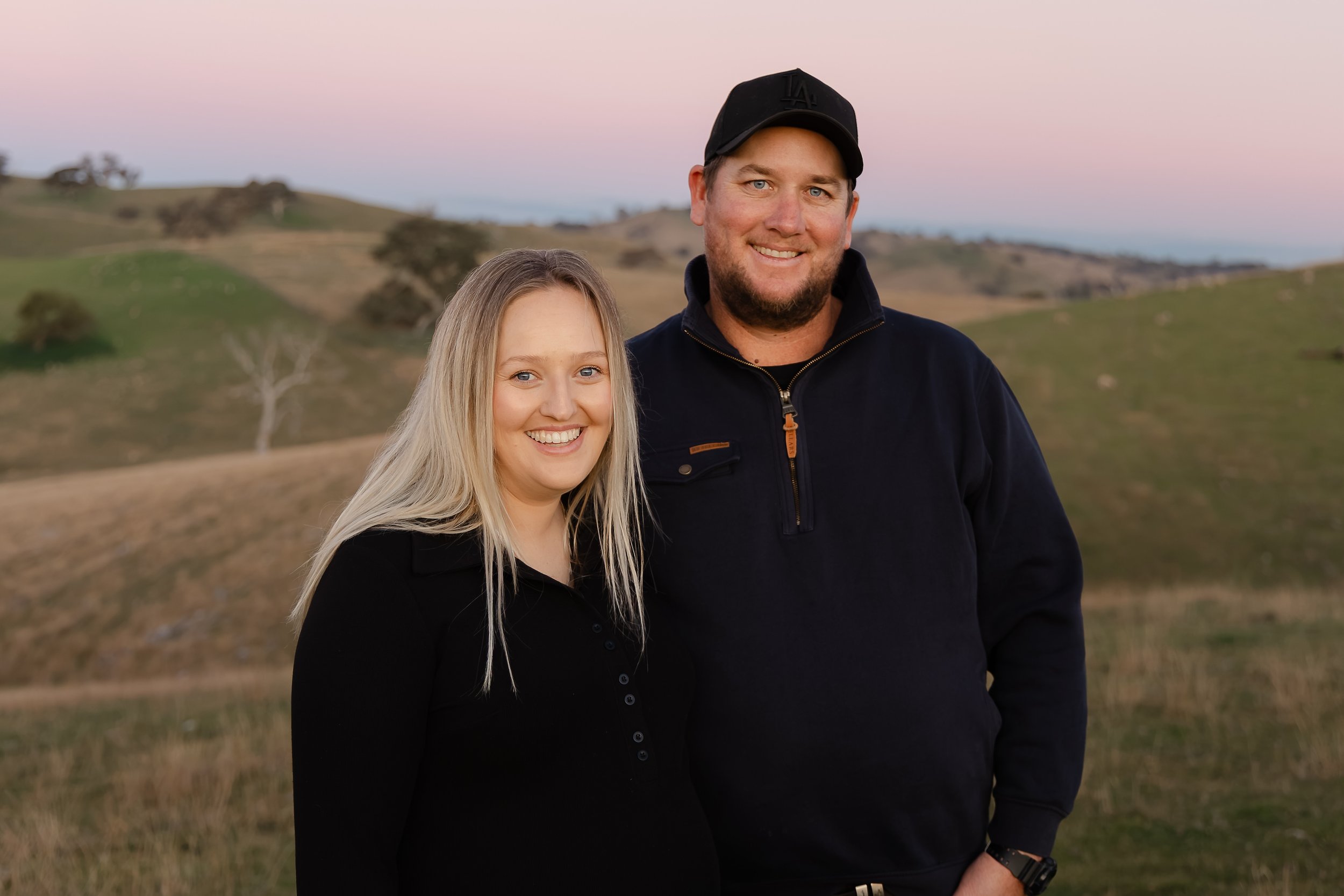 A young woman and a man standing outdoors in a grassy, hilly landscape during sunset, both smiling at the camera.