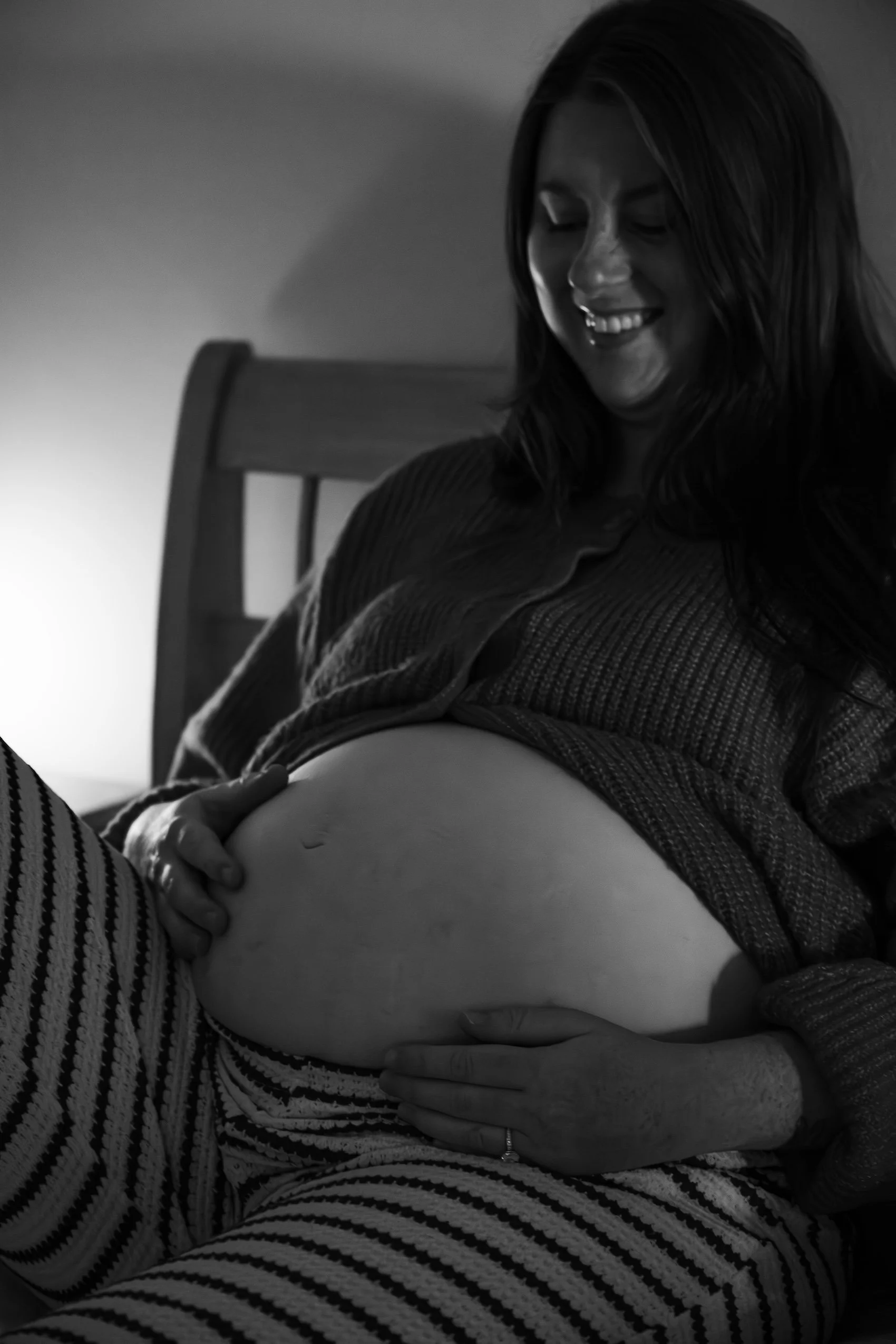A woman with dark hair smiling while looking down, wearing a striped long-sleeve shirt, sitting on a chair with a plain wall in the background. She is holding a pregnant belly with her hand.