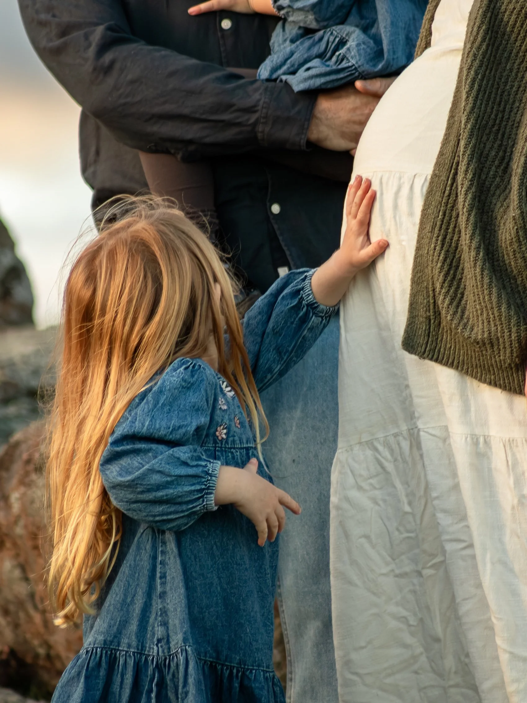 A young girl with long red hair wearing a denim jacket is touching a pregnant woman's belly, who is wearing a white dress and a brown cardigan. An adult man is standing nearby, holding a folded piece of cloth or scarf. The scene appears to be outdoor