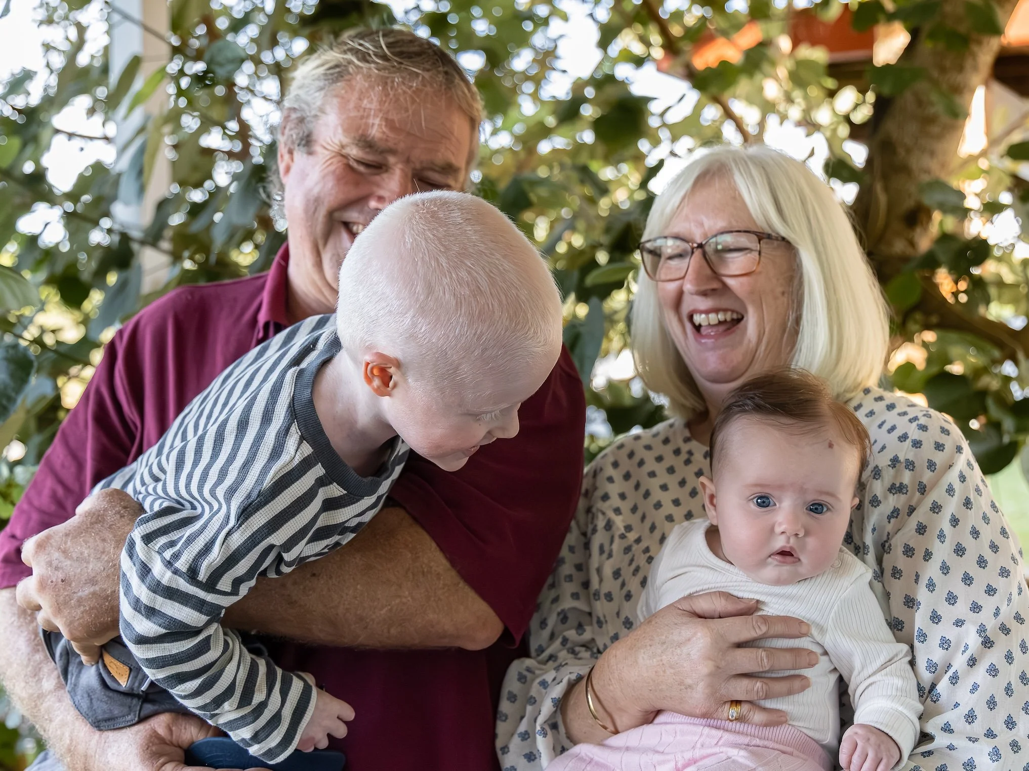 A happy multigenerational family, including elderly adults, a young child, and an infant, are gathered outdoors surrounded by trees, sharing a joyful moment together.
