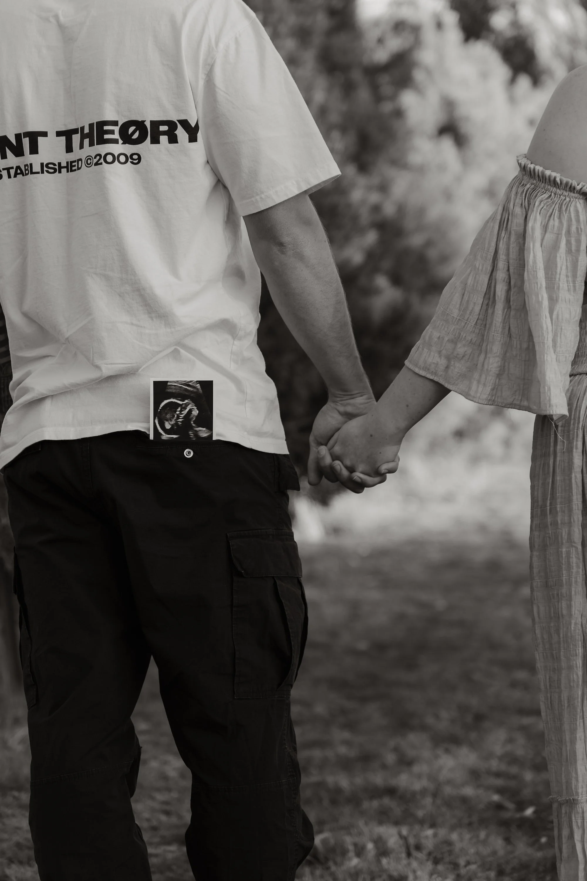 A man and woman holding hands, seen from the waist up, outdoors in a natural setting, both dressed casually, the man wearing a T-shirt with text and an ultrasound image, and the woman wearing a light-colored, ruffled sleeve top.
