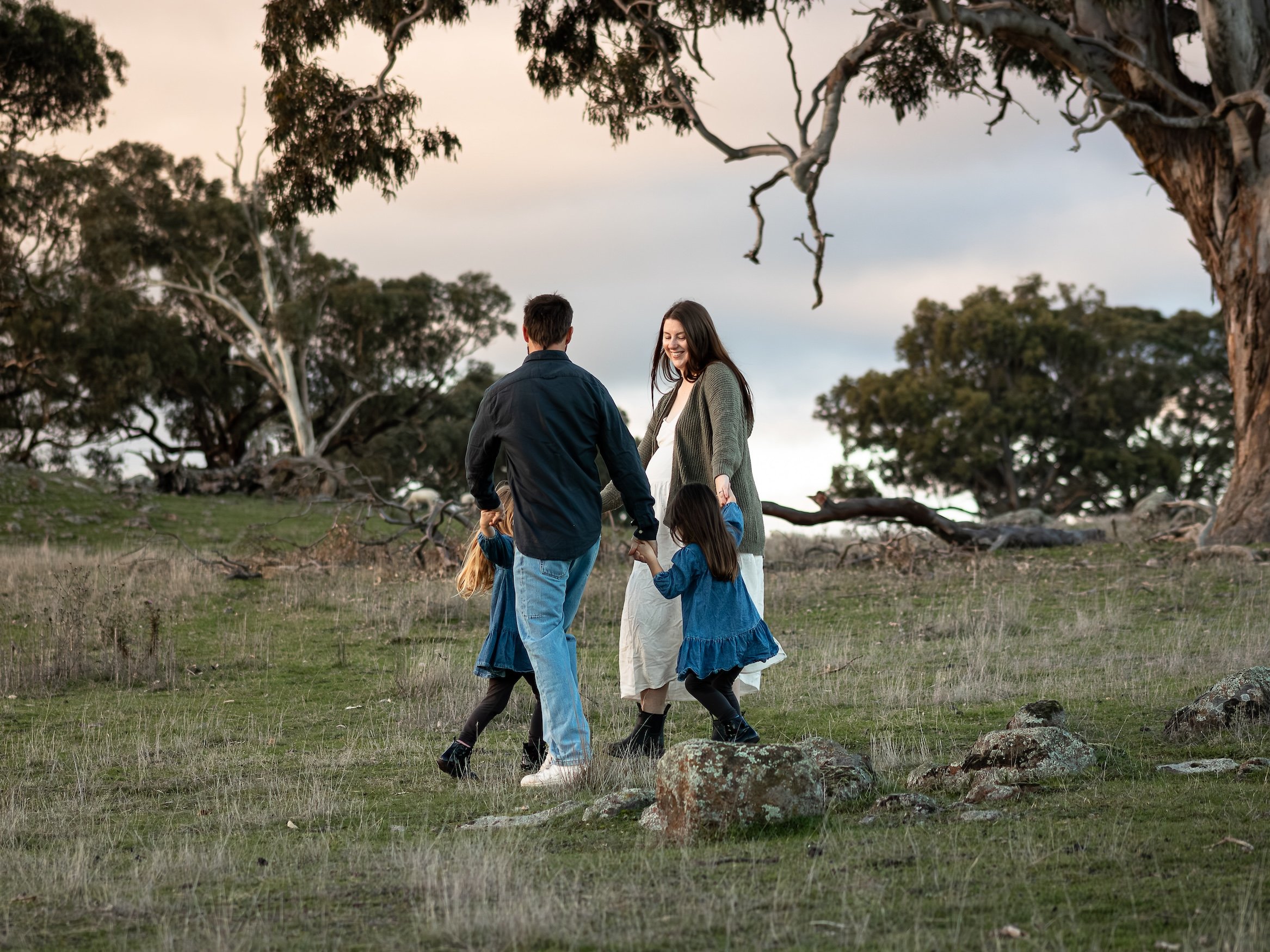 A family of five, including a mother, father, and three children, holding hands and walking in a grassy outdoor area with large trees around them as the sun sets or rises, casting a warm light.