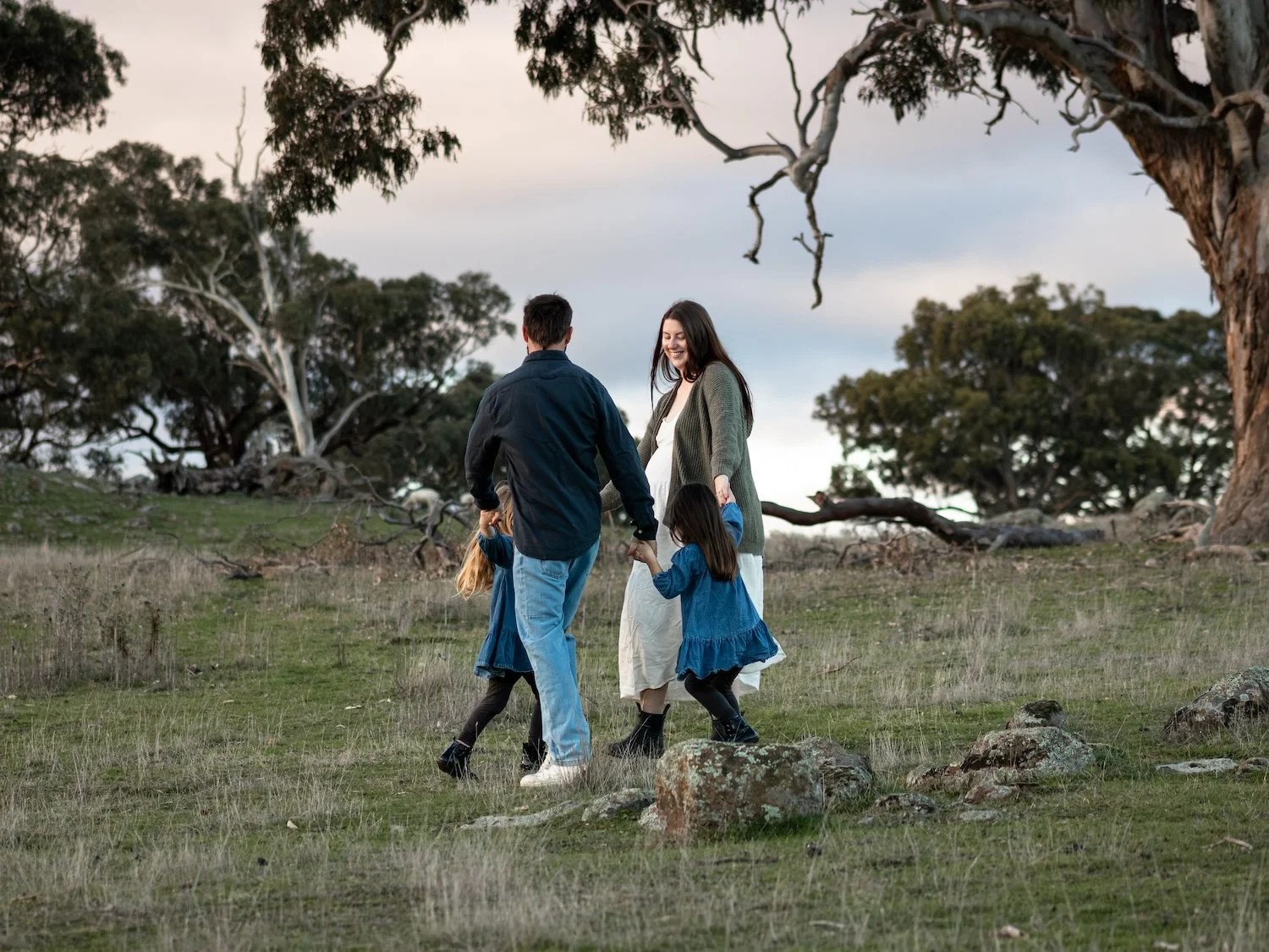 A family of four, including a pregnant woman, standing with two young children in a grassy field with trees and cloudy sky in the background during sunset.