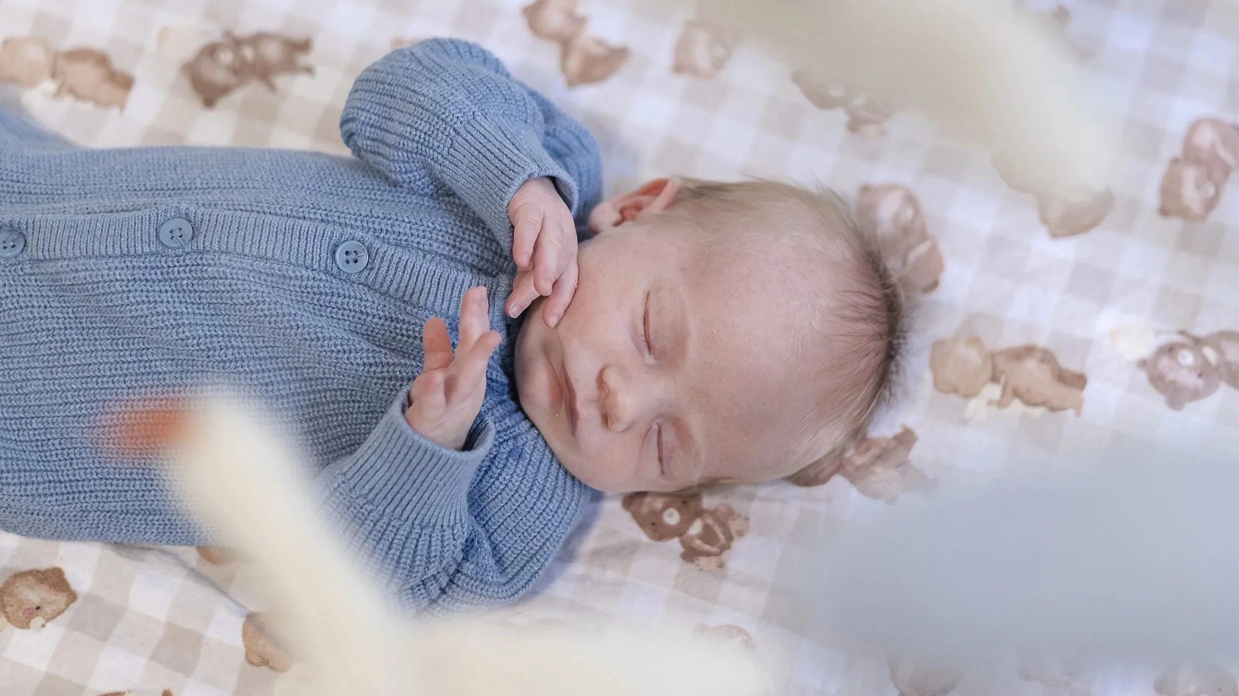 Newborn baby lying peacefully in his cot wearing a blue knit onesie during an in-home lifestyle newborn photoshoot in Canberra, captured through his cot mobile by Monique Patmore Photography.