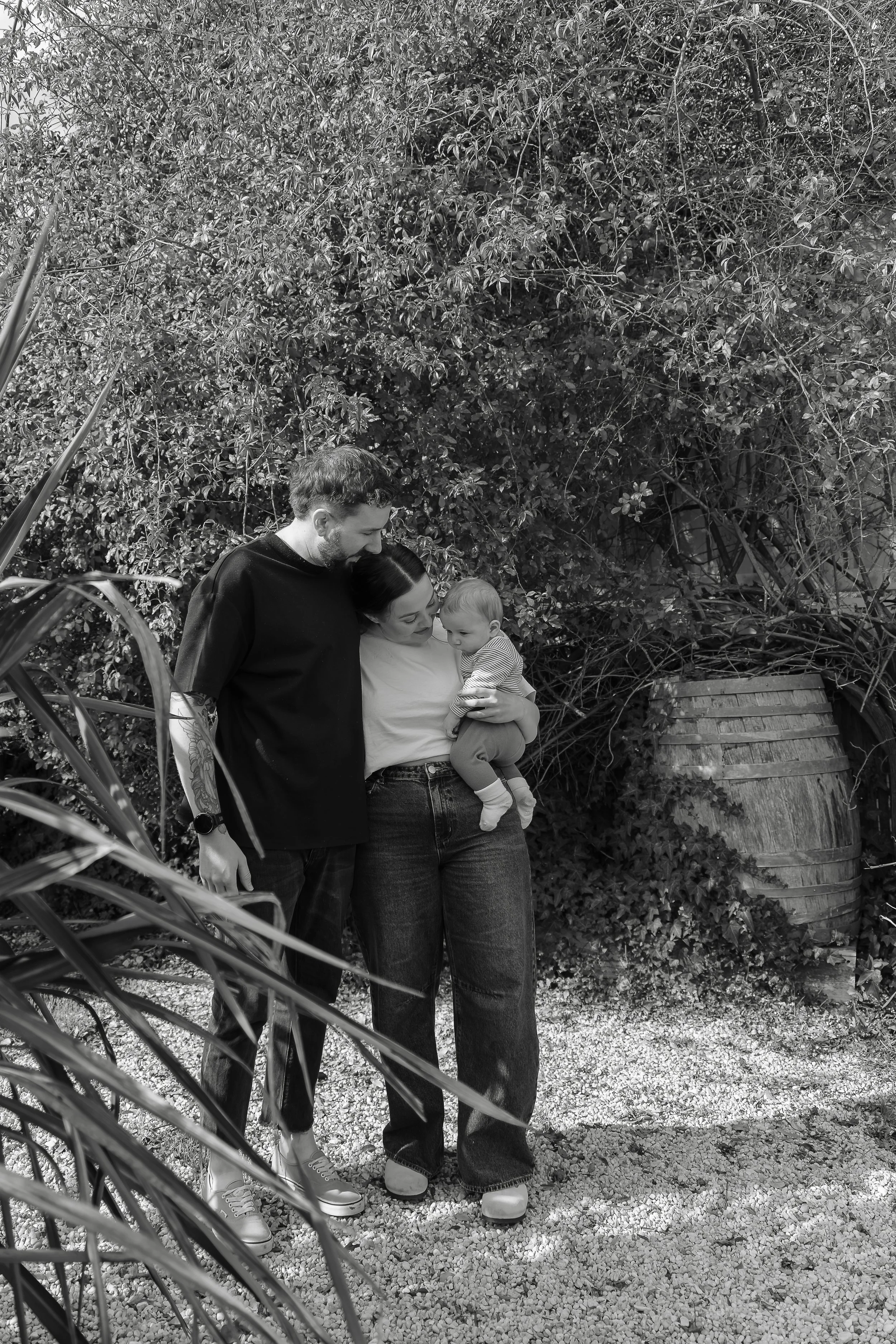 A family of three standing outdoors in front of dense foliage, with a woman holding a small child, a man leaning towards them, all engaged in a moment together.