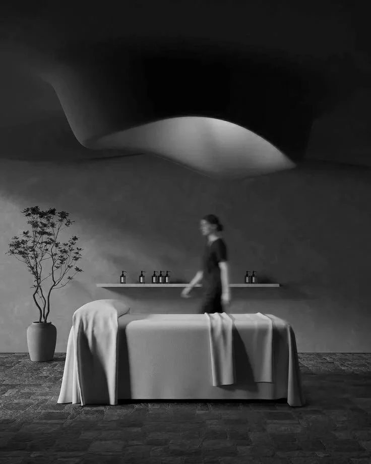 A woman walking past a massage table covered with a cloth in a modern room. In the background, there are bottles on a shelf and a potted plant. The ceiling has an unconventional, curved design.