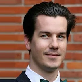 A man with dark hair and fair skin, wearing a dark blazer over a light shirt, standing in front of a brick wall.