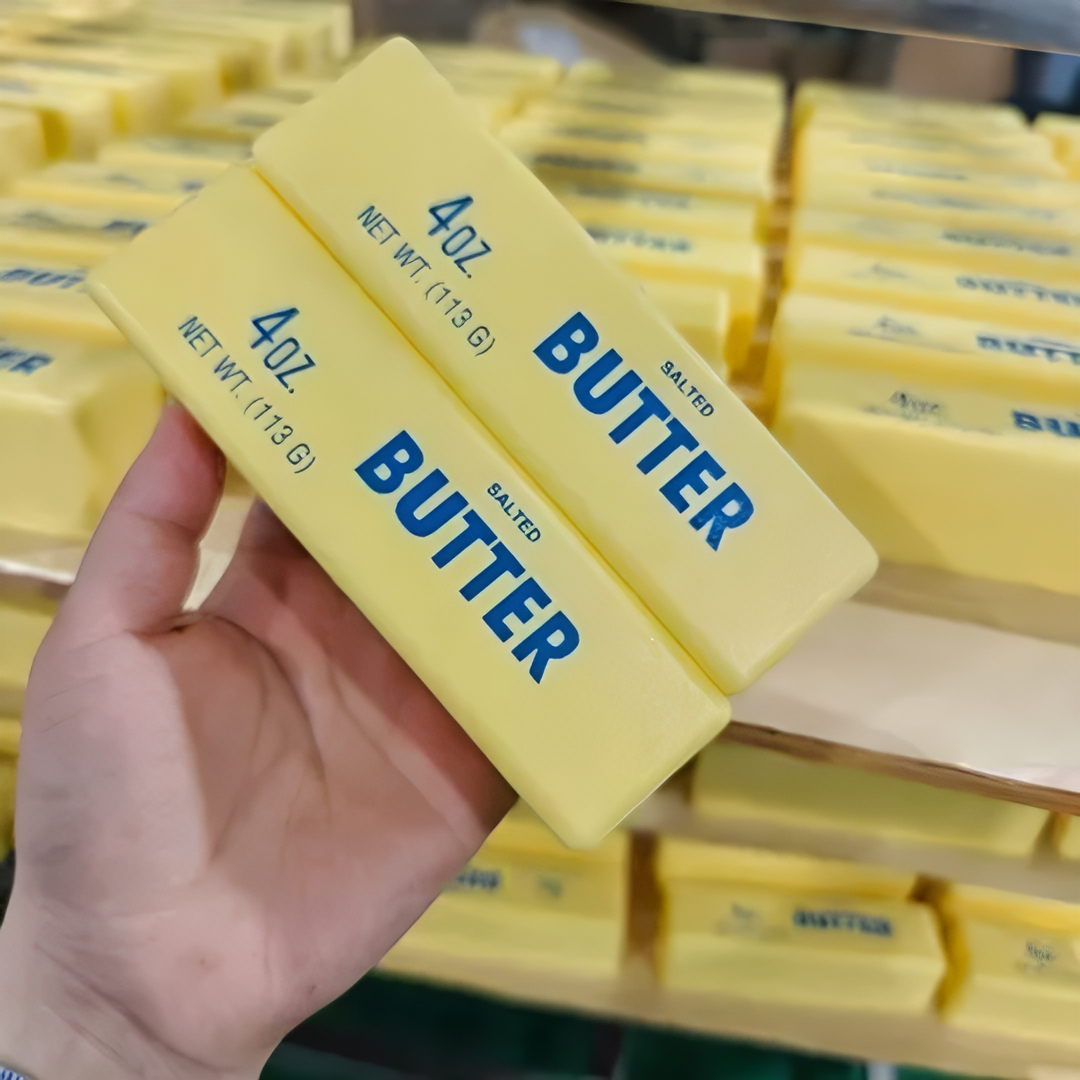 A person holding two sticks of salted butter in a grocery store. The butter is in yellow packaging with blue text that reads 'Butter.'