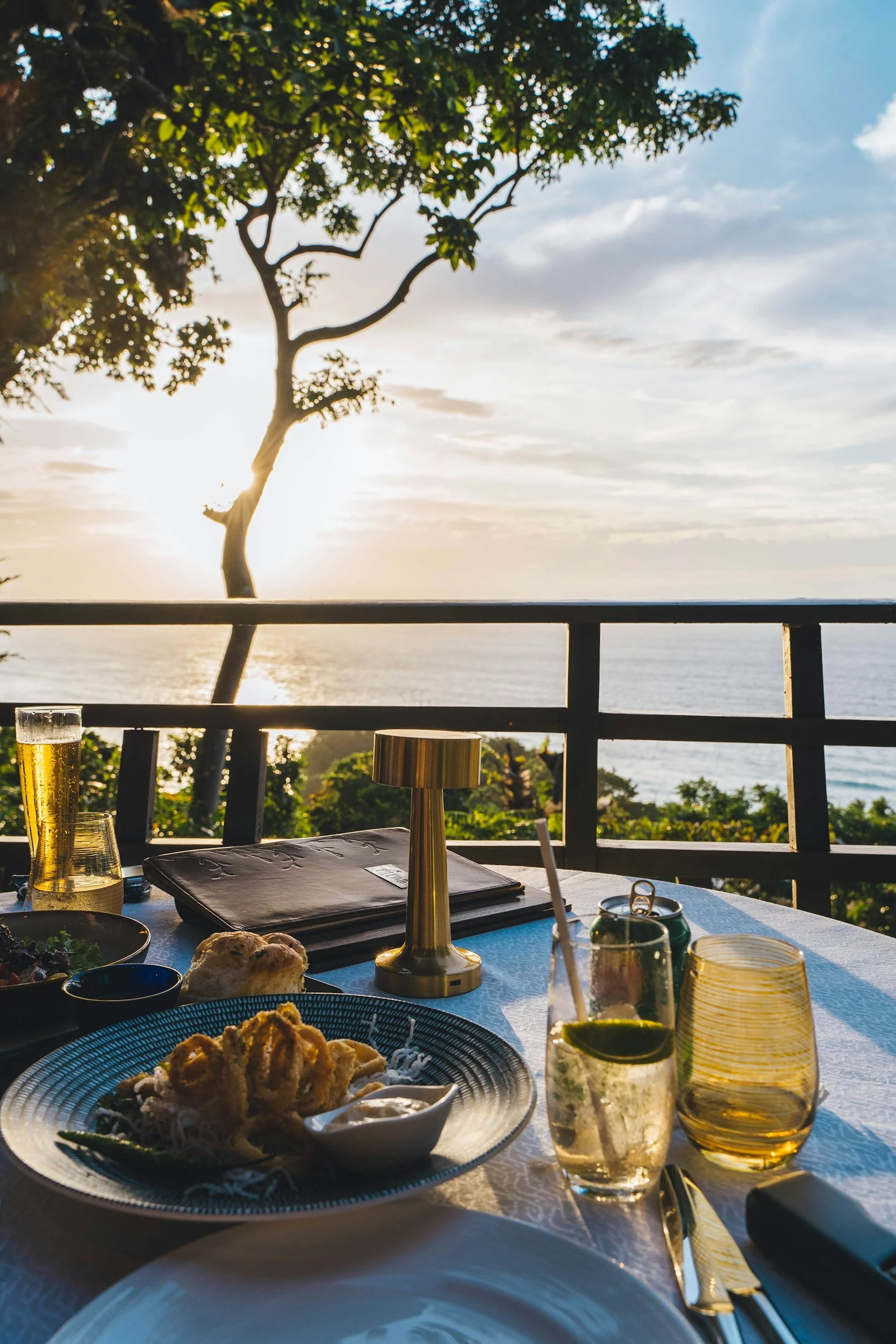 A table with plates of food, drinks, and utensils on a balcony overlooking the ocean, with a tree in the background and the sun setting.