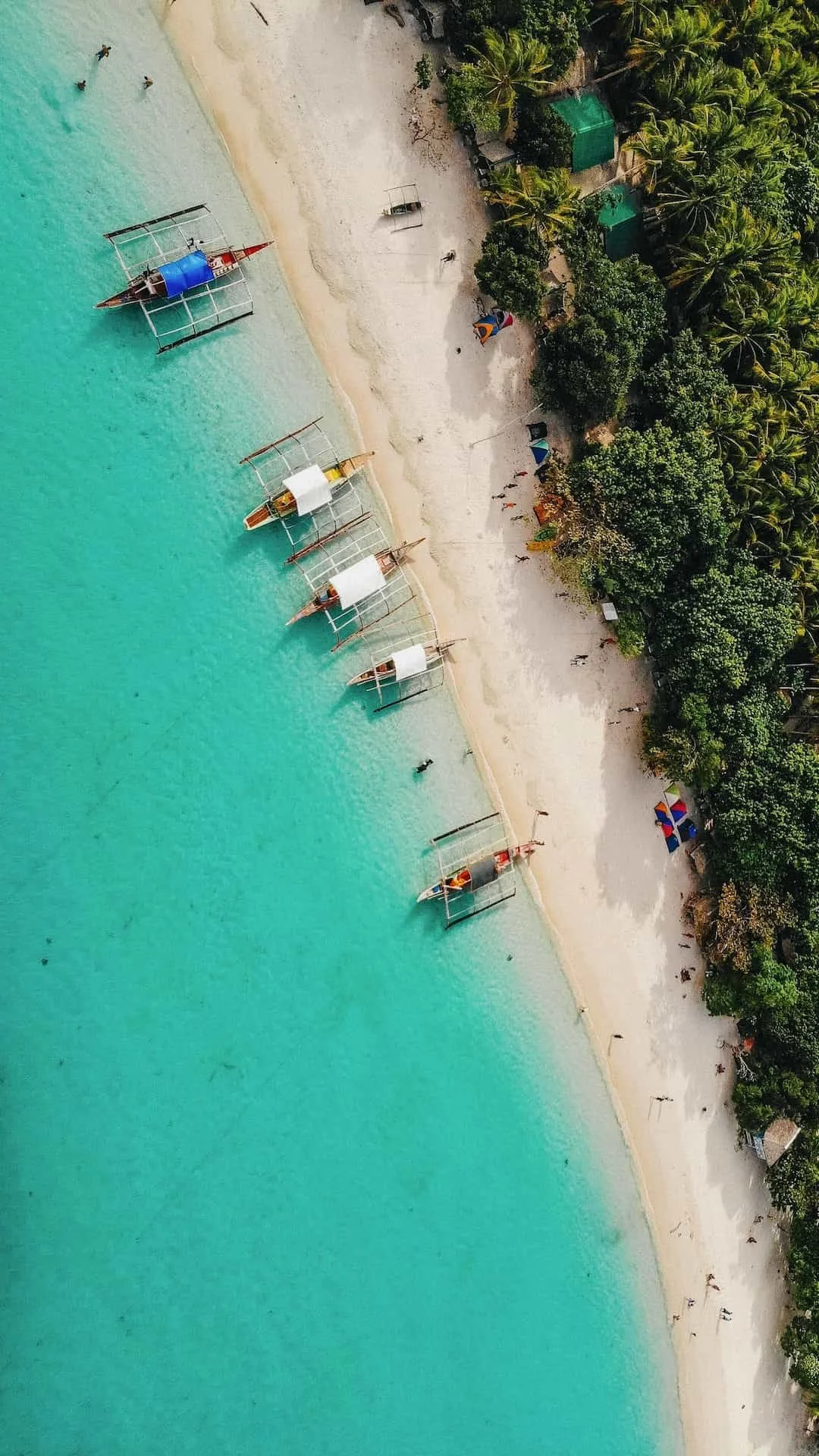 Aerial view of a tropical beach with turquoise water, boats with white roofs anchored near the shore, and lush green trees along the sandy coastline.