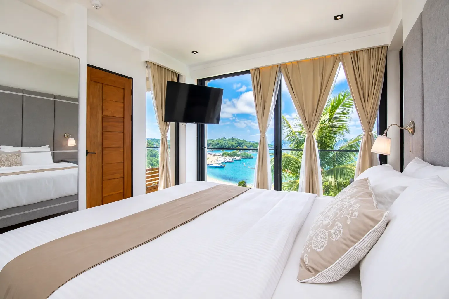 Modern hotel room with a large bed, bedside lamps, and an ocean view through floor-to-ceiling windows with beige curtains, overlooking a marina with boats and green landscape.