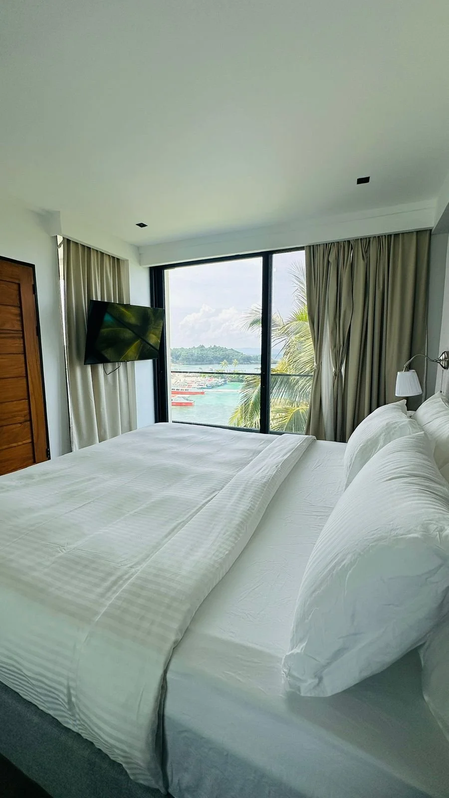 A hotel room with a white bed, large glass window showing a view of the ocean and palm trees, beige curtains, a wall-mounted TV, and a lamp beside the bed.