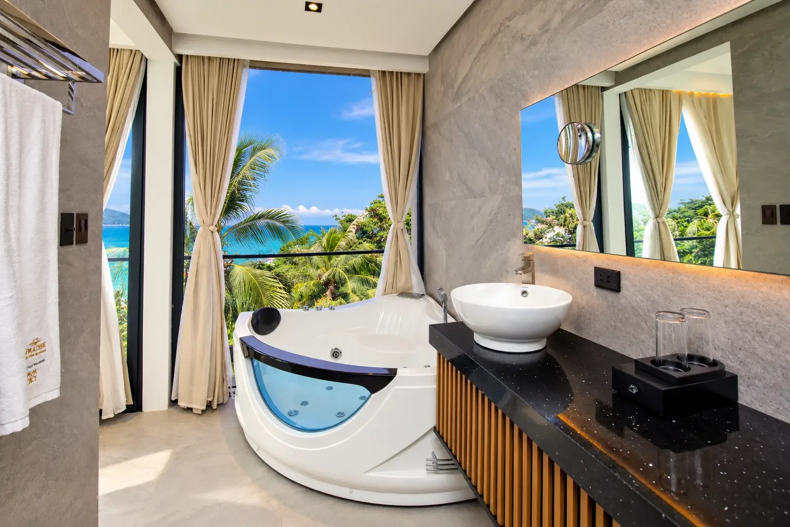 Luxury bathroom with a jacuzzi tub overlooking a tropical landscape and the ocean, large window with beige curtains, a black granite countertop with a vessel sink, mirror, and glassware.