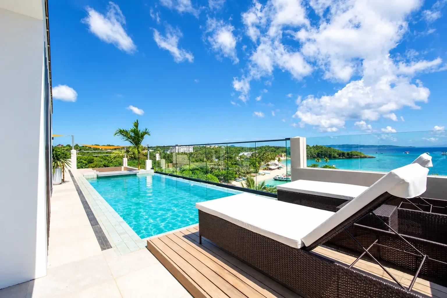 Rooftop pool area with lounge chairs overlooking a tropical beach and ocean under a blue sky with clouds.