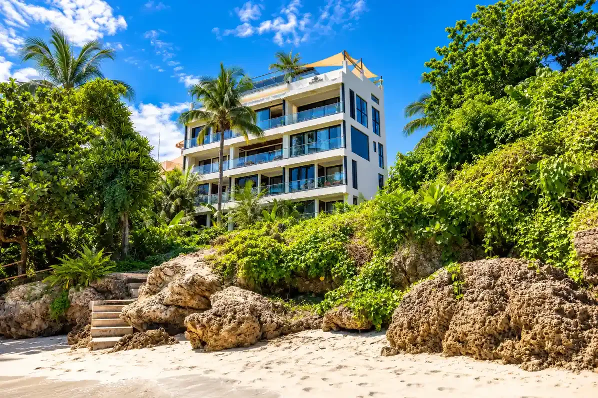 Modern white multi-story beachfront building with glass balconies, surrounded by palm trees and rocks on sandy beach under blue sky
