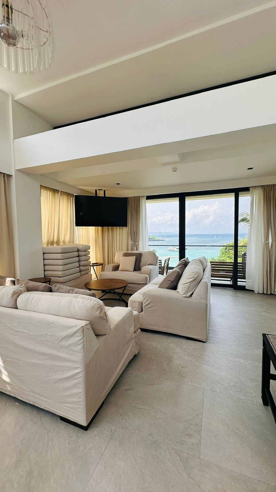 Living room with white sofas, a round wooden coffee table, beige curtains, a wall-mounted TV, and large glass sliding doors showing an ocean view with a balcony and blue sky.