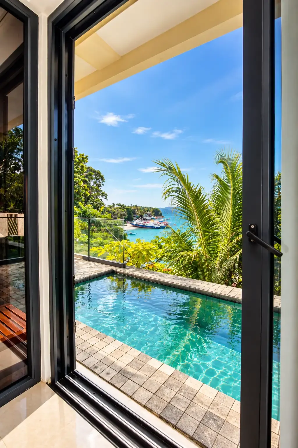 View from an open sliding glass door showing a small swimming pool on a balcony with tropical plants, a hillside with lush trees, and a body of water with boats under a clear blue sky.