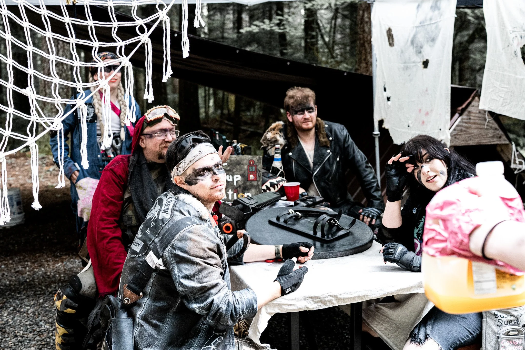 Group of people dressed in post-apocalyptic or zombie costumes sitting and standing around a table outdoors in a wooded area, with some wearing face paint, masks, and accessories, under a structure with hanging fabric or sheets.