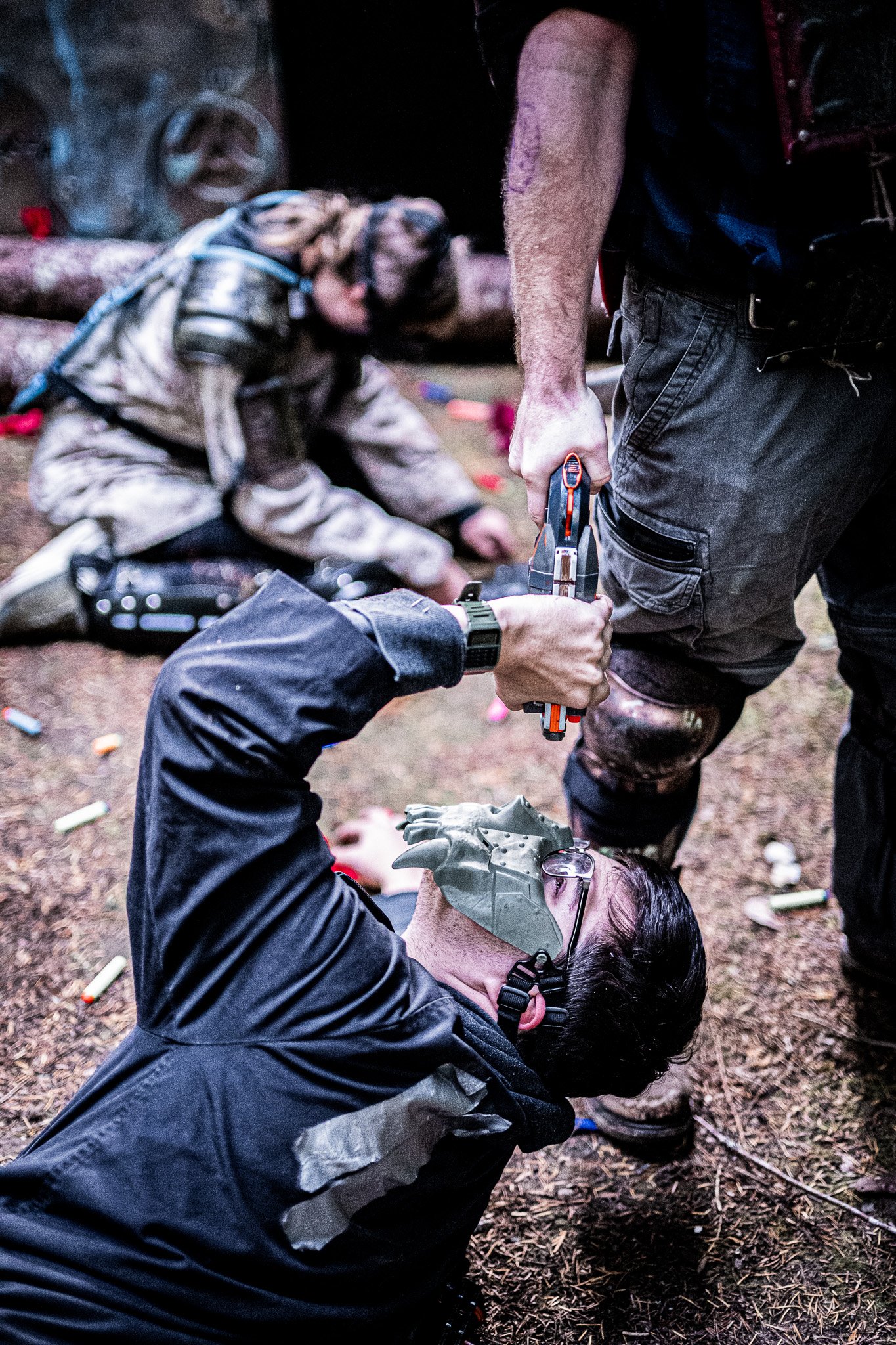 A person lying on the ground with a makeshift eye patch over their eye, wearing glasses, and another person kneeling beside them, holding a firearm and tending to their injury. The scene appears chaotic with empty shells scattered on the ground.