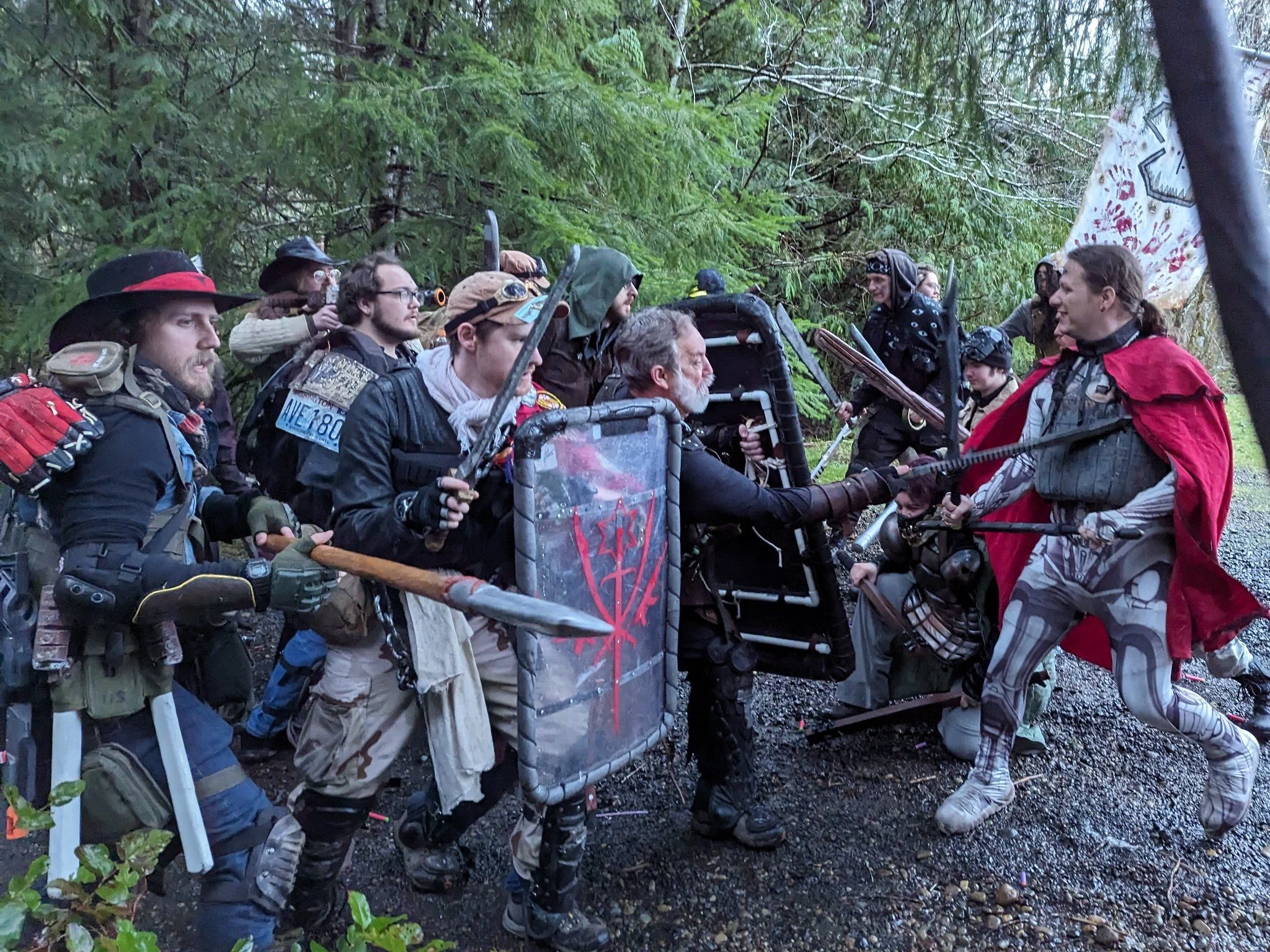 A group of people dressed in fantasy or medieval costumes stand in a dense forest, holding weapons and shields, appearing to participate in a live-action role-playing event.