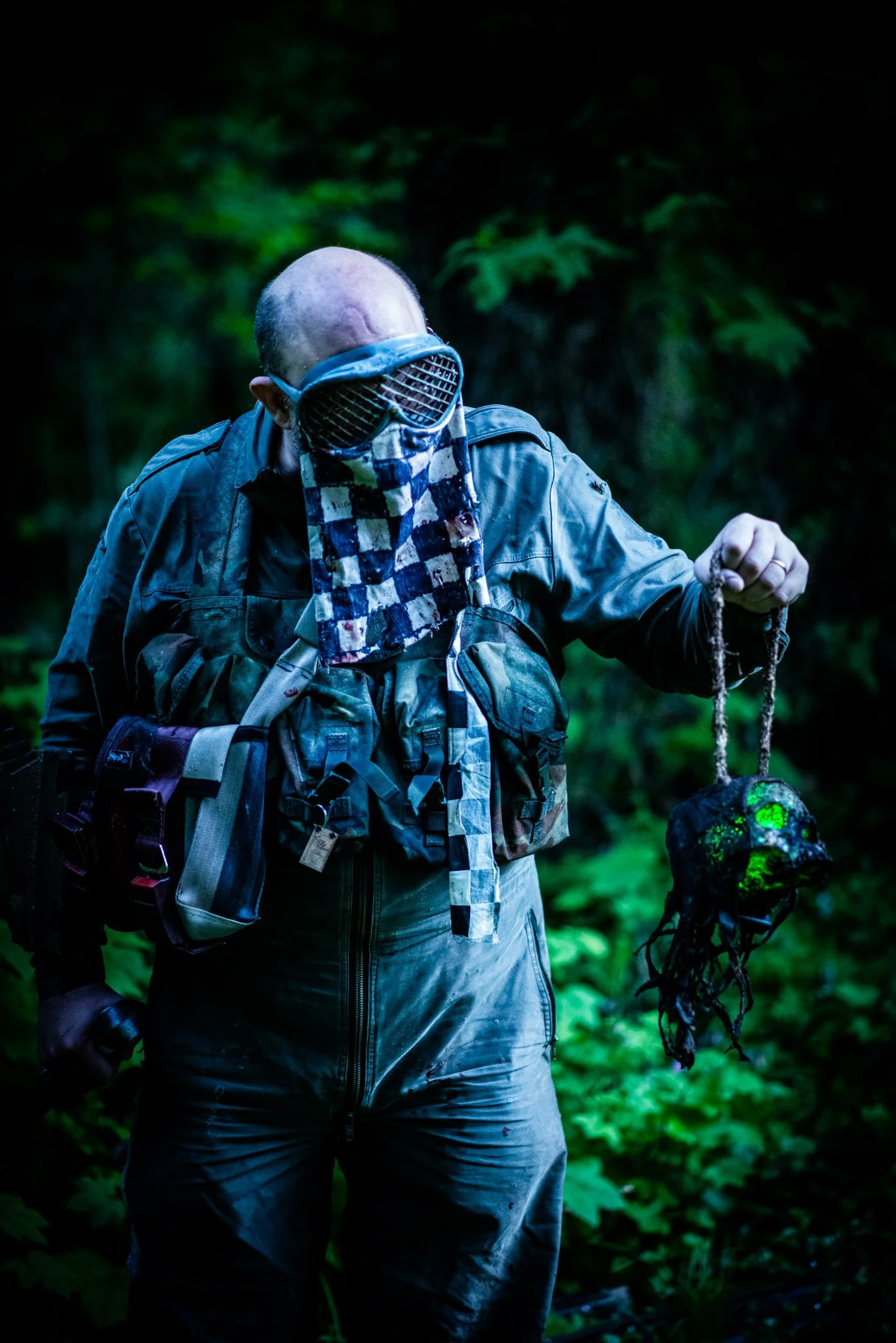 A person in outdoor gear, wearing a jacket, gloves, and camouflage face covering, holding a luminescent object or artifact in a forest at night.