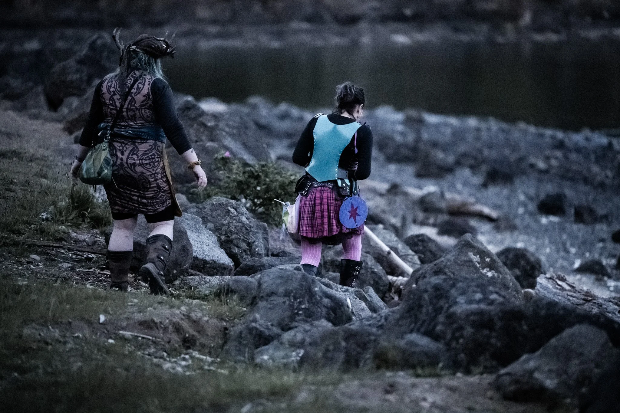 Two women walking along a rocky shoreline at dusk, one wearing a patterned dress and the other wearing a plaid skirt with a blue vest and holding a walking stick.