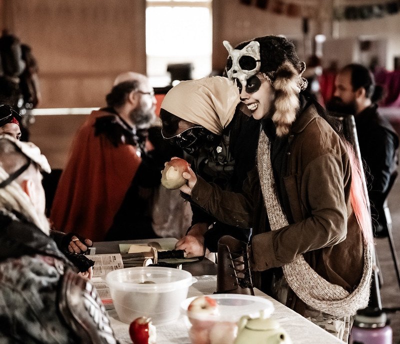 People in costume at a market or festival, engaging with vendors at a booth.