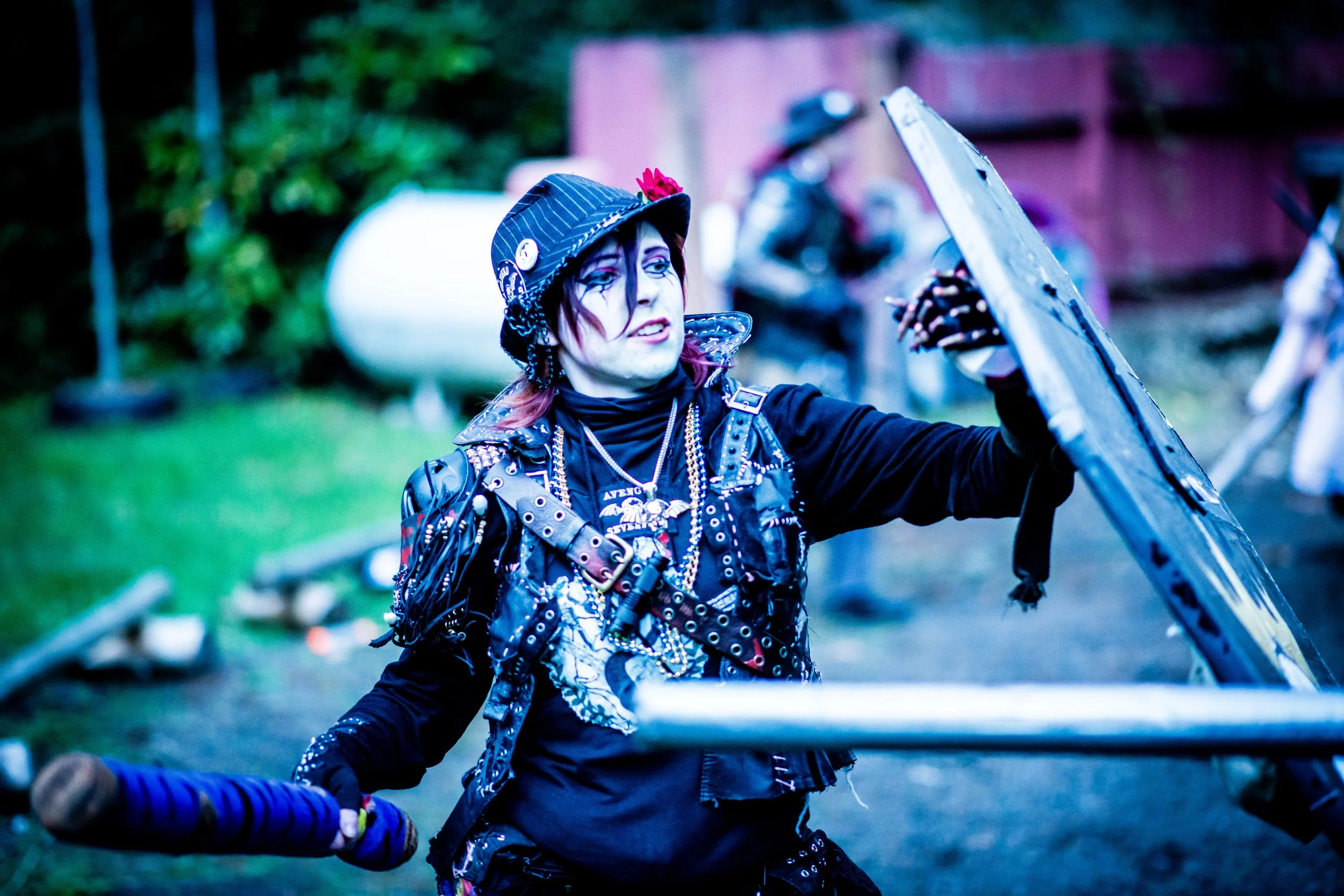 Person dressed in punk style costume with face makeup, wearing a black and white striped hat with buttons, holding a large sword, standing outdoors with blurred background of trees and other figures.