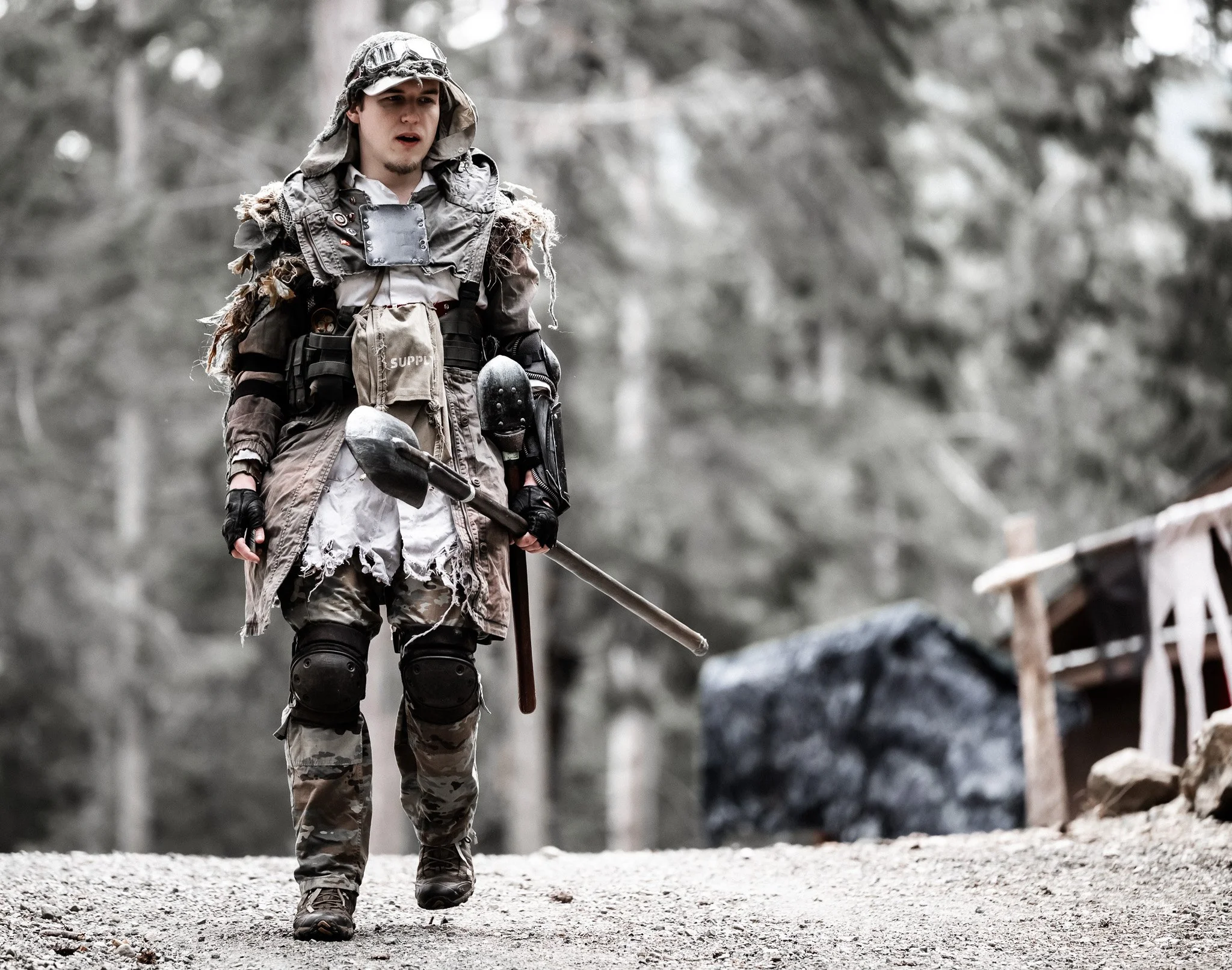 A person dressed in military or tactical gear walking outdoors in a forested area, holding a shovel, with a serious expression.