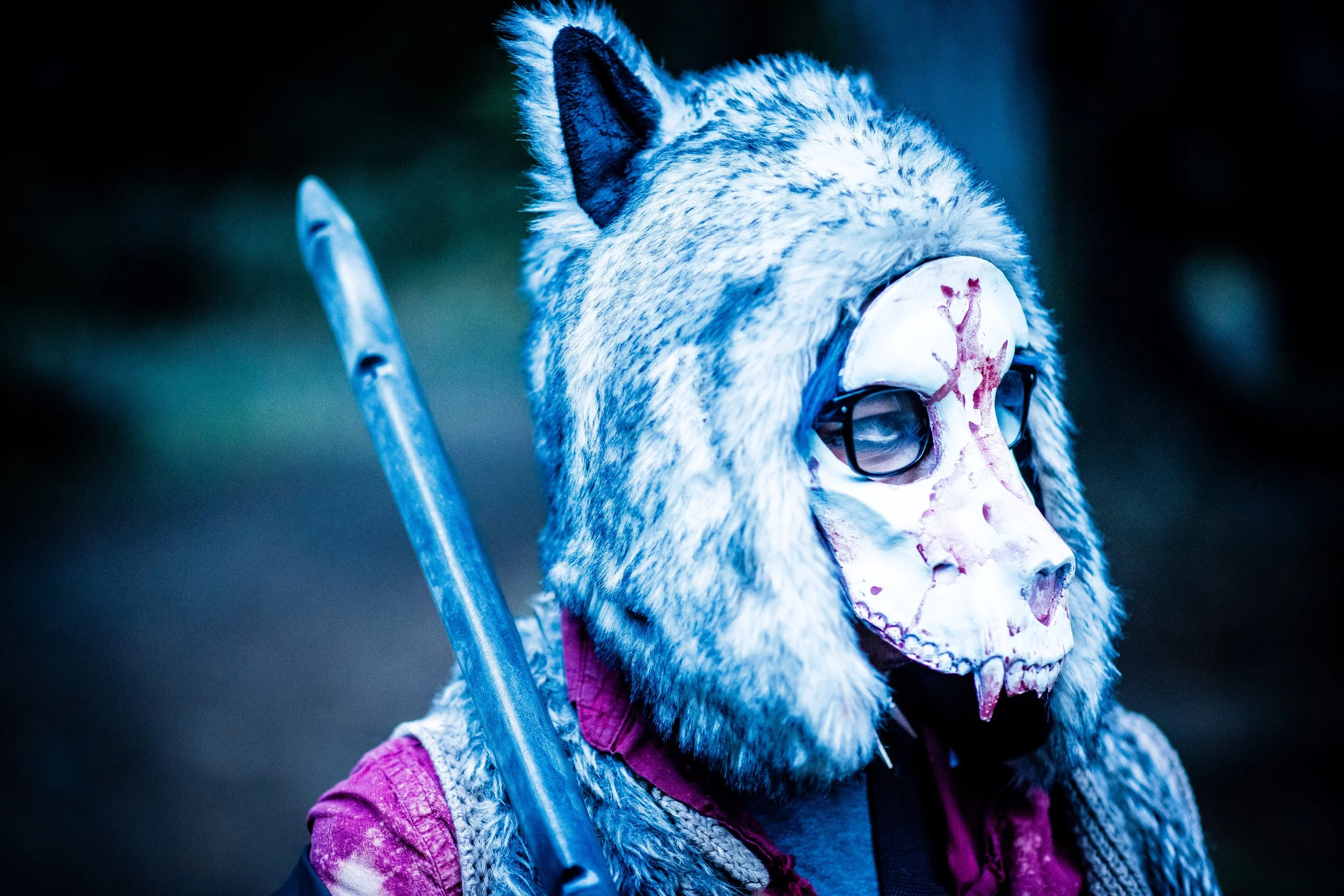 Person wearing a wolf mask with glasses, purple jacket, and a firearm, standing outdoors in low light.