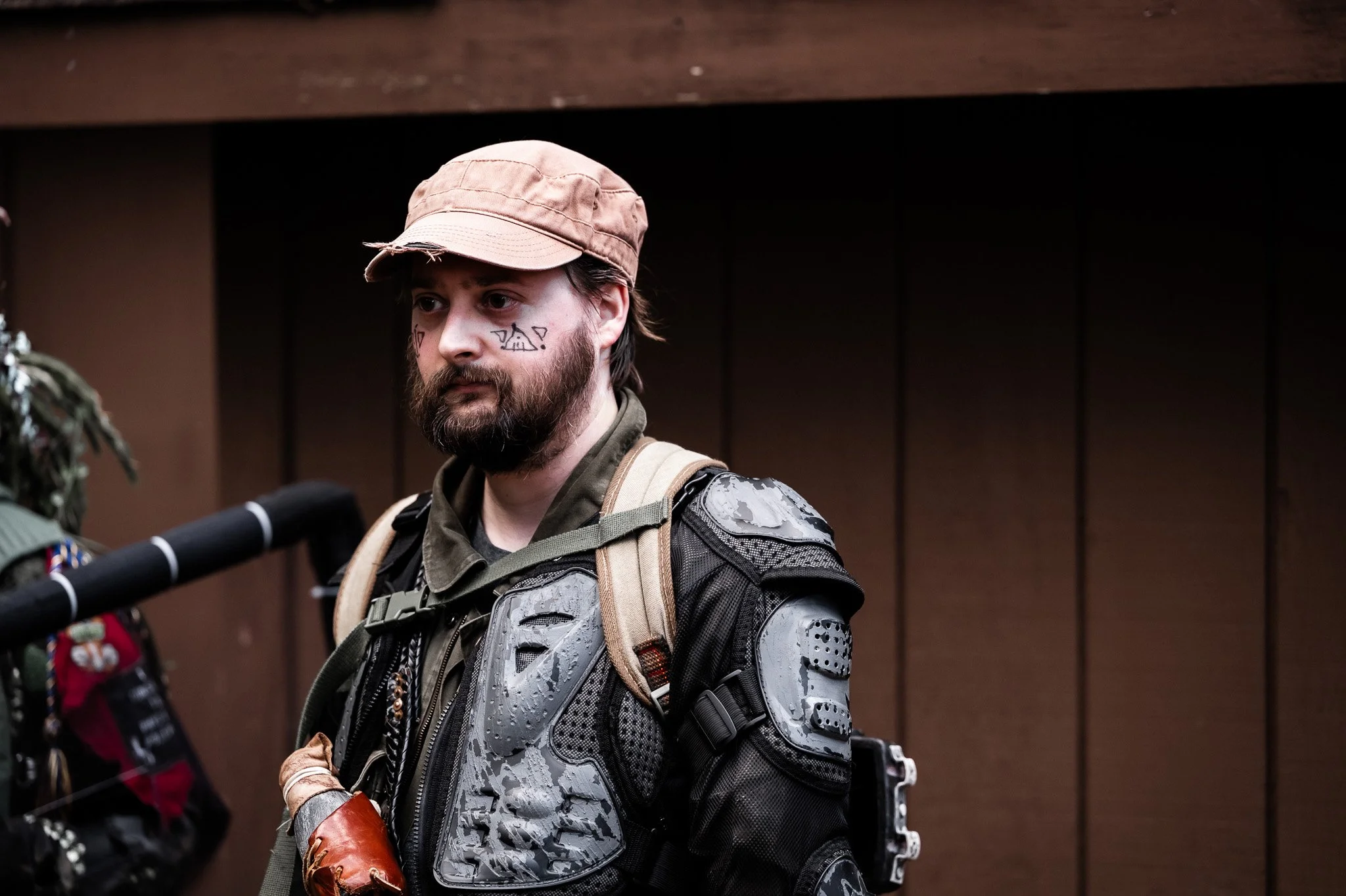 A man with a beard, face tattoos, and a pink cap, dressed in tactical gear with painted armor, standing indoors in front of a brown wall.