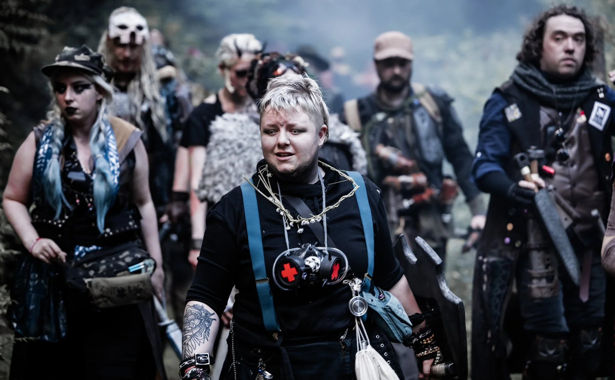 Group of people dressed in post-apocalyptic or zombie-themed costumes, walking through a wooded area with smoke, with a young man in the foreground wearing a black shirt, backpack, and fake injuries.