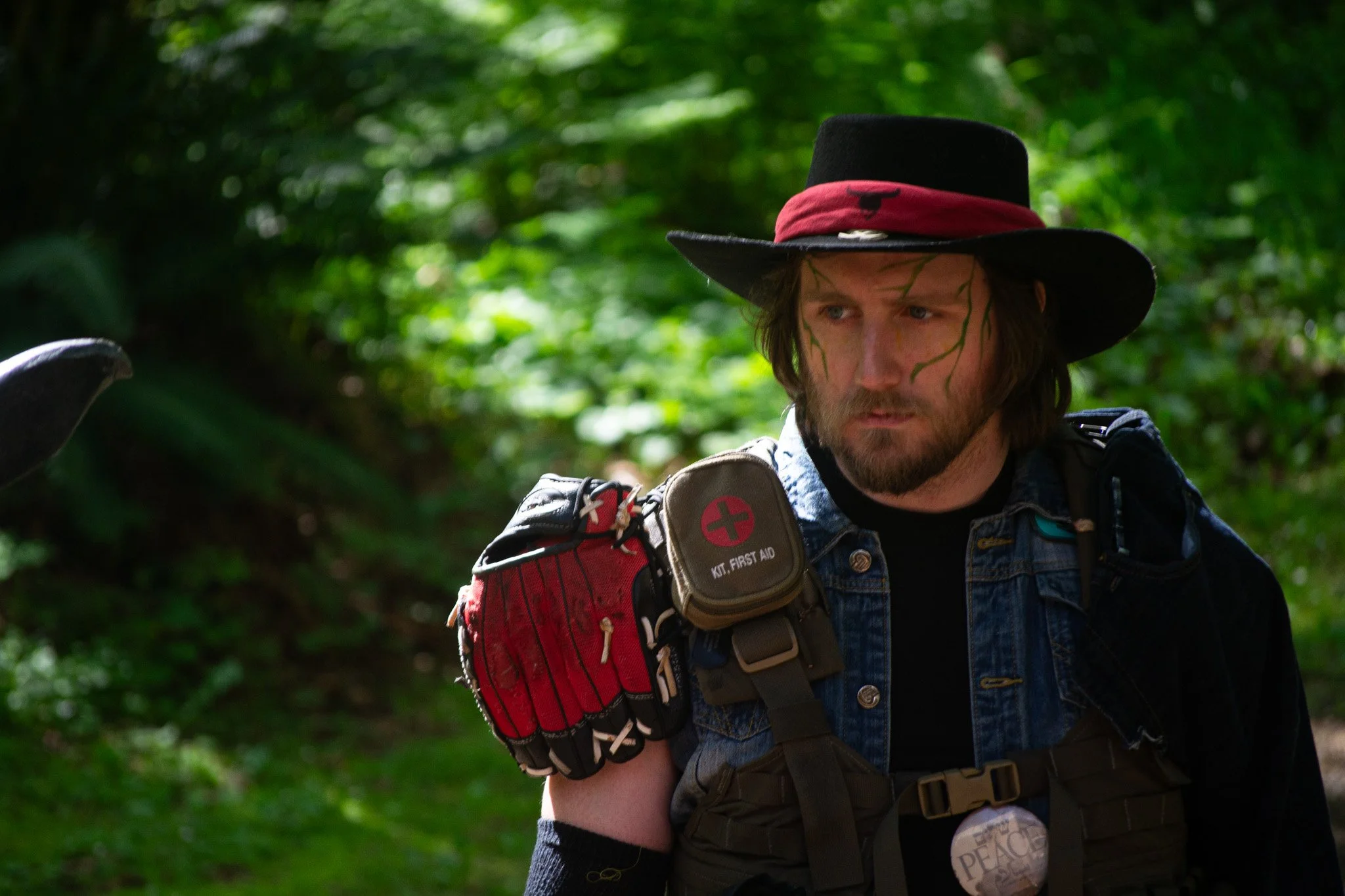A man in a black hat with a red band and a bird emblem, wearing camouflage face paint, a denim vest, and carrying a red backpack with first aid kit, walking in a lush green forest.