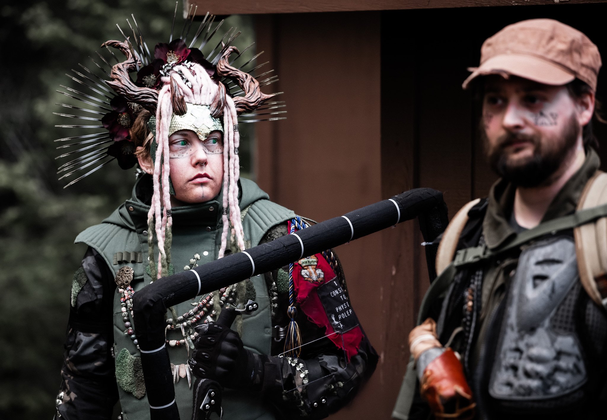 Person wearing an elaborate headdress with feathers, flowers, and dreadlocks, holding a pole, with another person in military gear beside them.