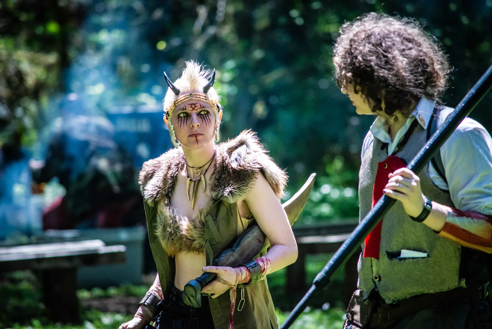 Two people in elaborate costumes and face paint, one with a headdress and fur, the other with curly hair and a patterned vest, stand in a lush forest.