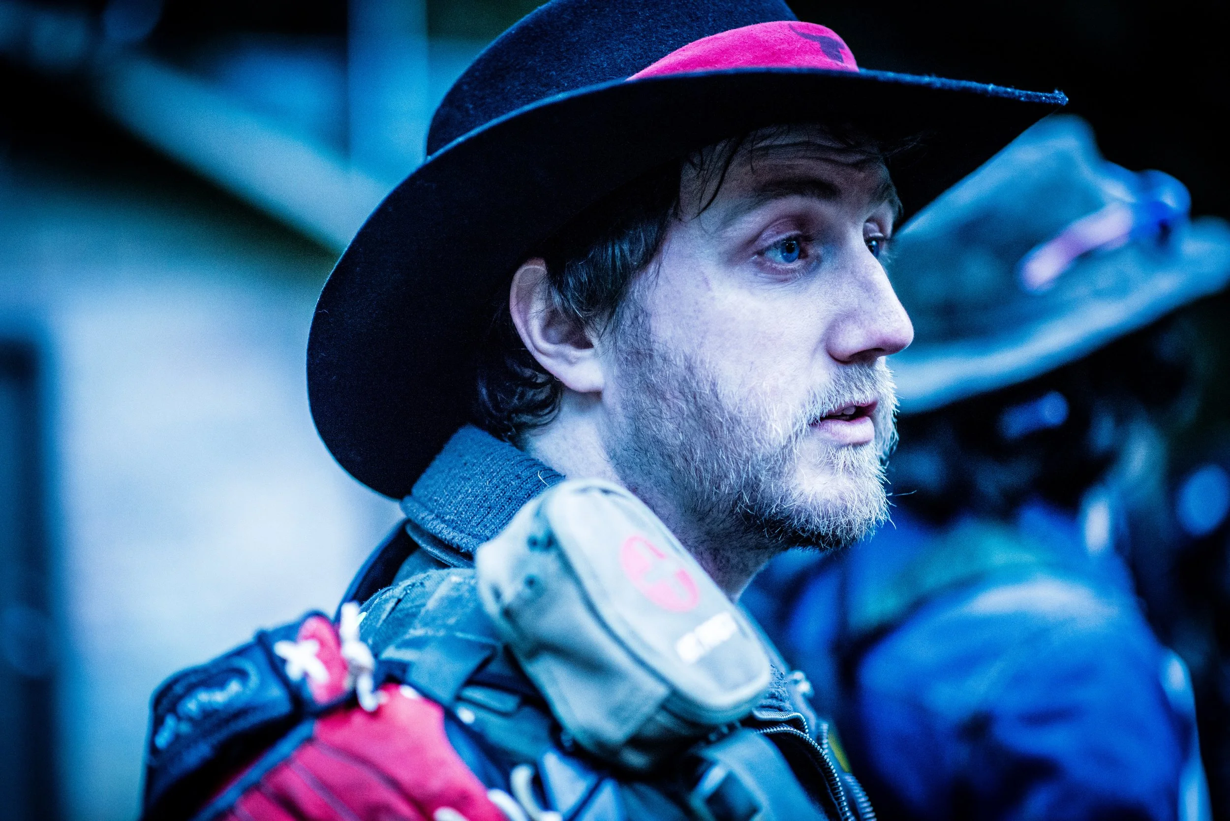 Close-up of a man with blue eyes, a beard, and long dark hair, wearing a black and pink hat and a jacket with a gray and pink pattern, holding a camera or phone, with a blurred background.