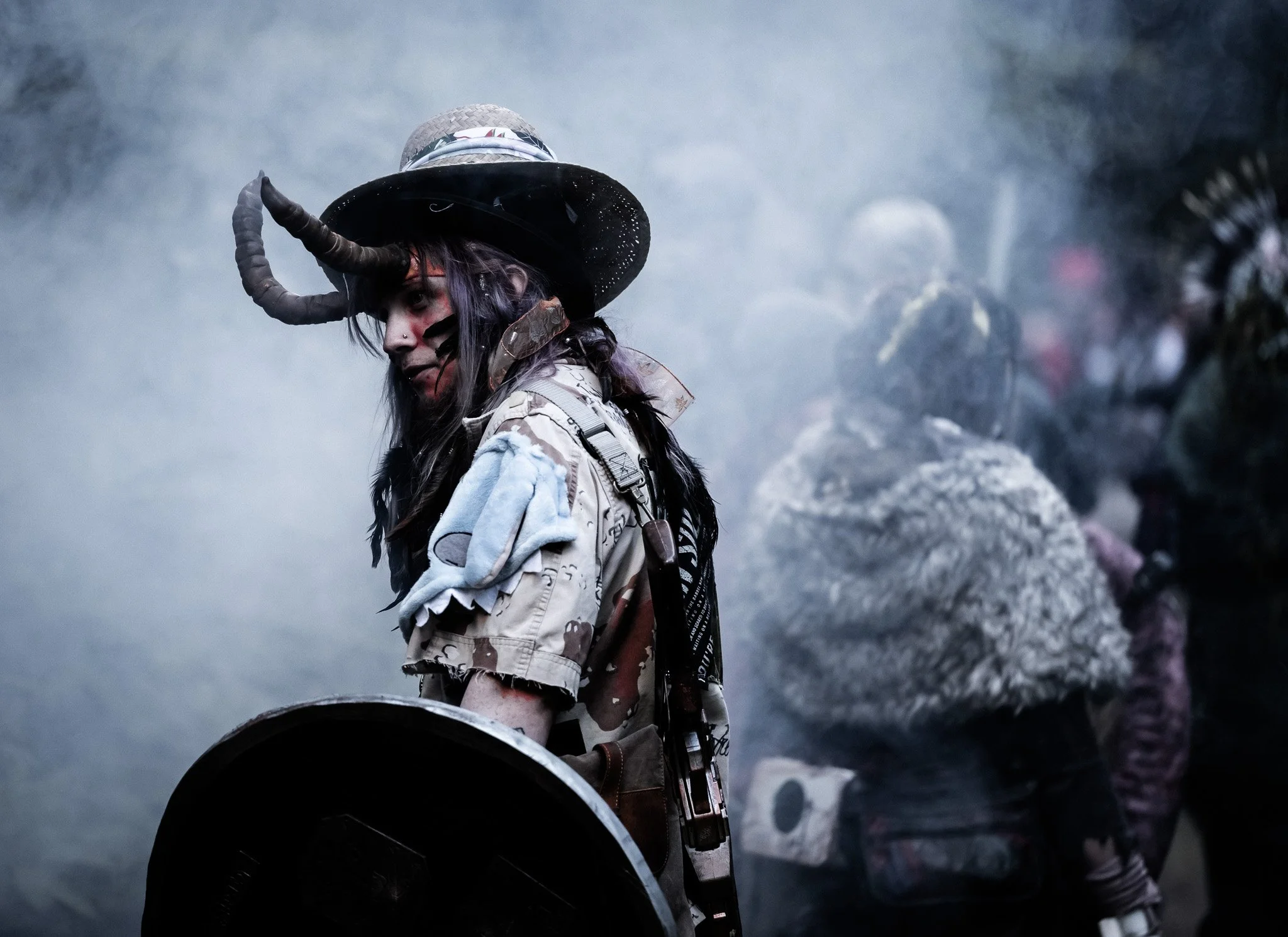A person with long dark hair, painted face, wearing a cowboy hat, camouflage jacket, and backpack, standing among smoke and flames, holding a shield or a similar object in a smoky, chaotic environment.