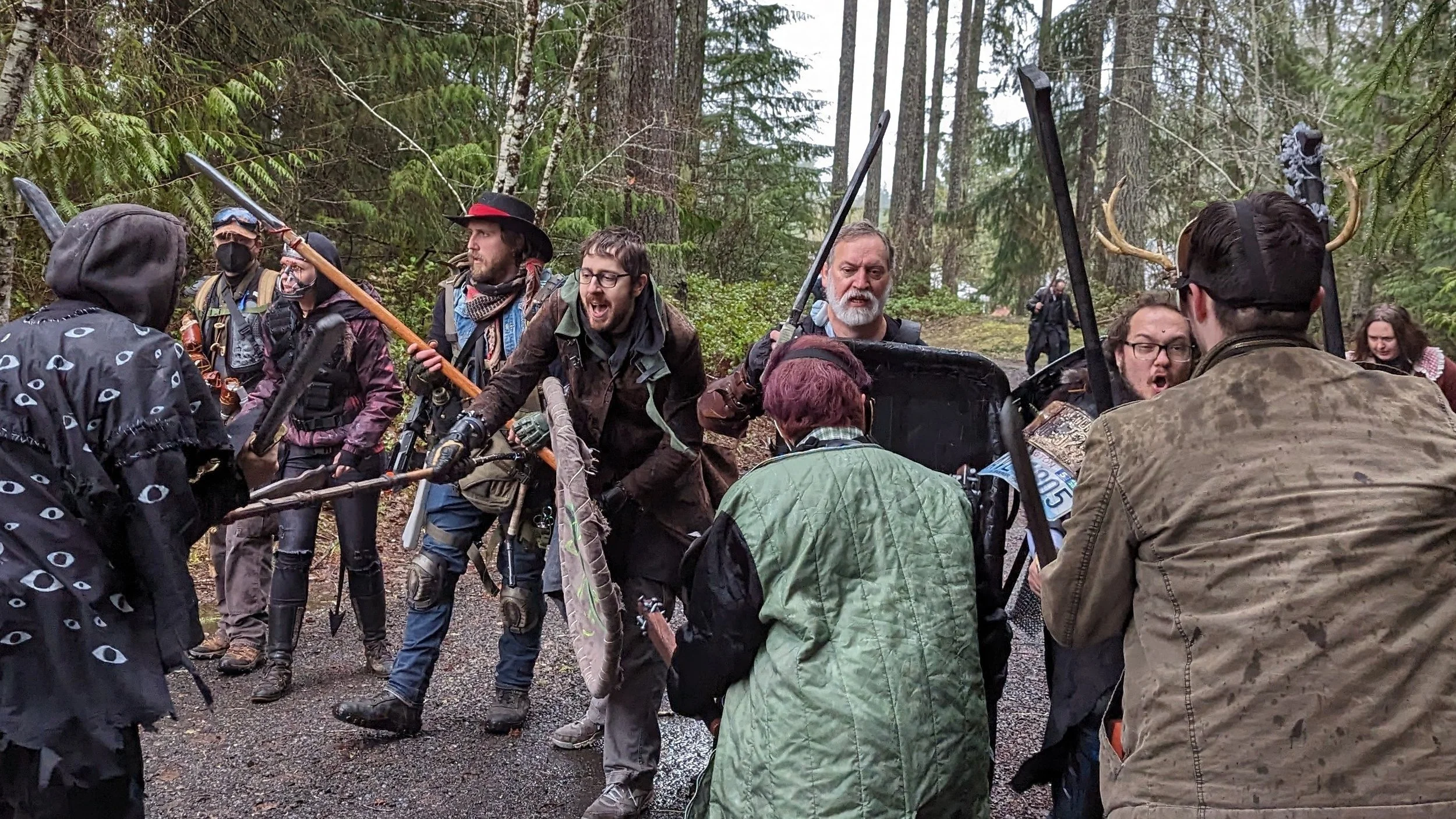 Group of people in forest, some holding weapons, appearing to confront each other, with trees in background