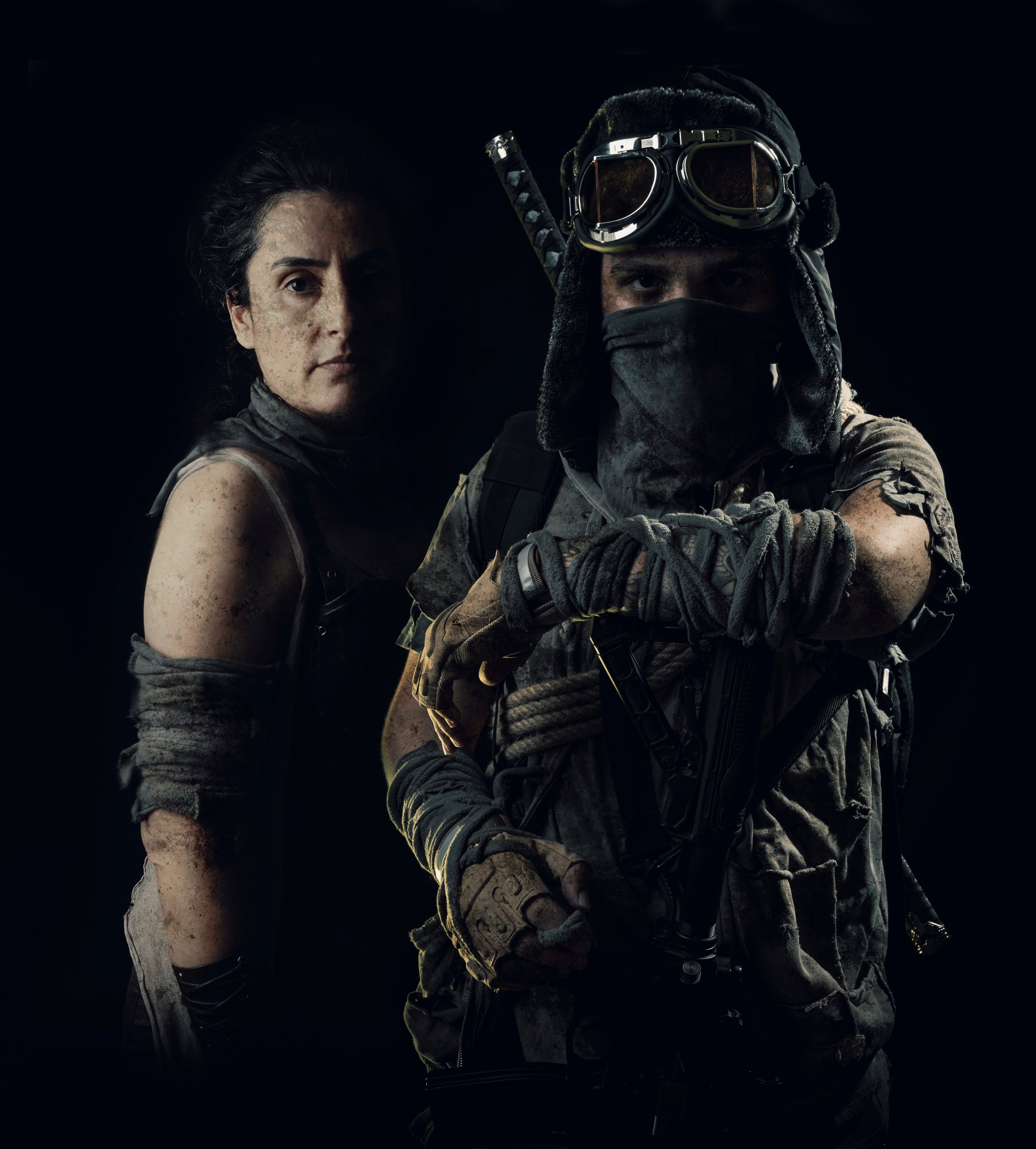 Two soldiers in combat gear, one with face covered and goggles, the other with dirt on face and a serious expression, against a dark background.
